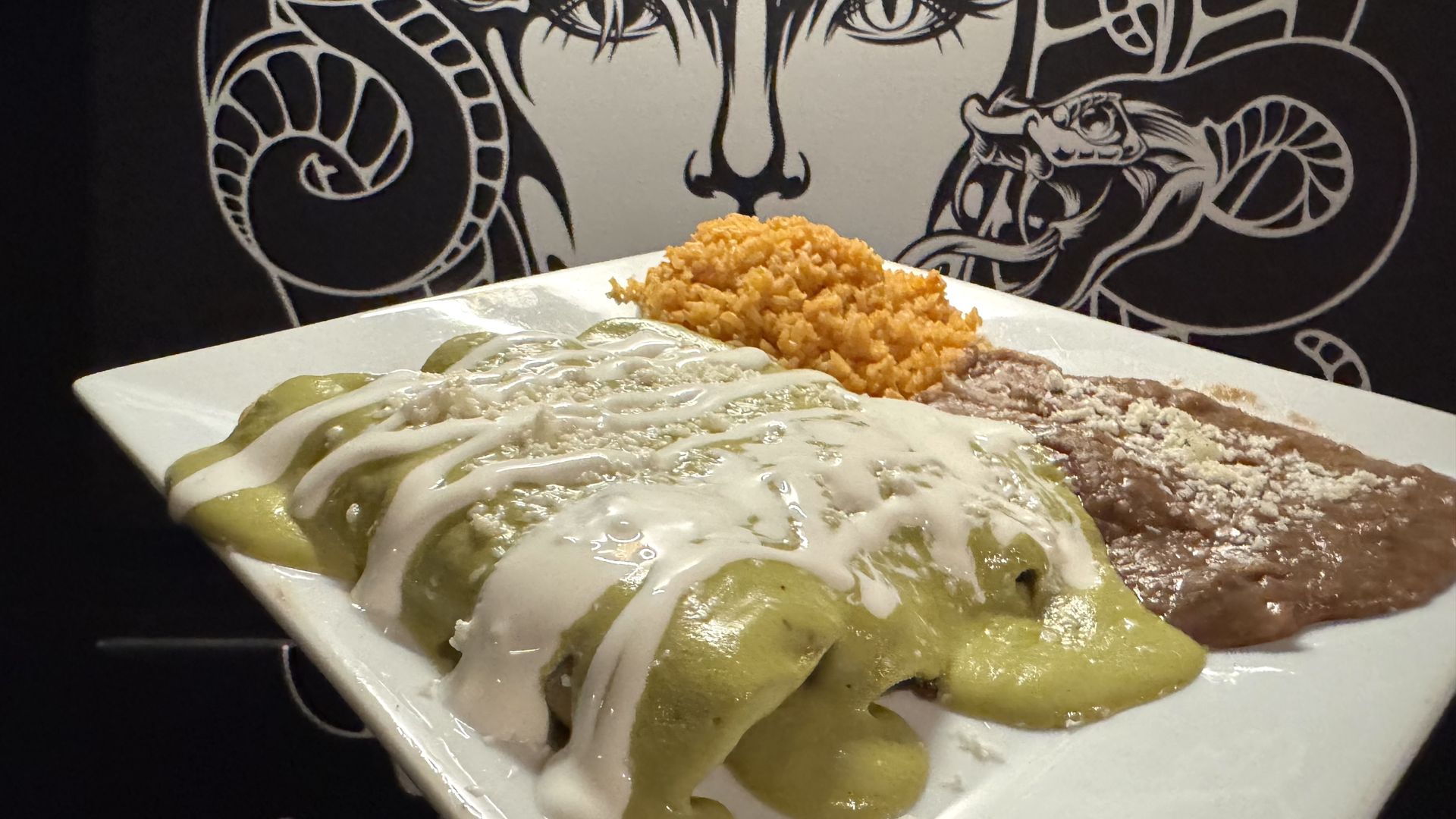 A plate of enchiladas topped with green sauce and crema, served with Mexican rice and refried beans, in front of a black-and-white mural with snake and face designs.