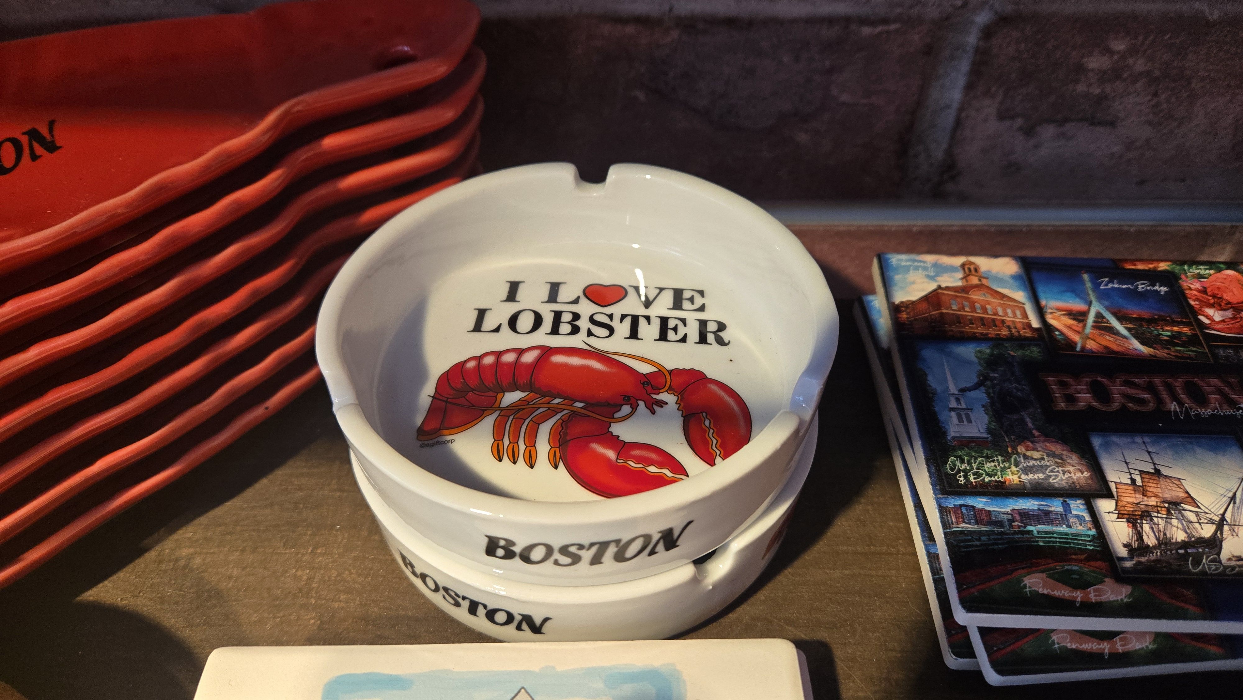 White ashtrays stacked, one shows red lobster and text "I ♥ LOBSTER" with "BOSTON" on side. Red plates stacked nearby and Boston souvenir magnets with landmarks.