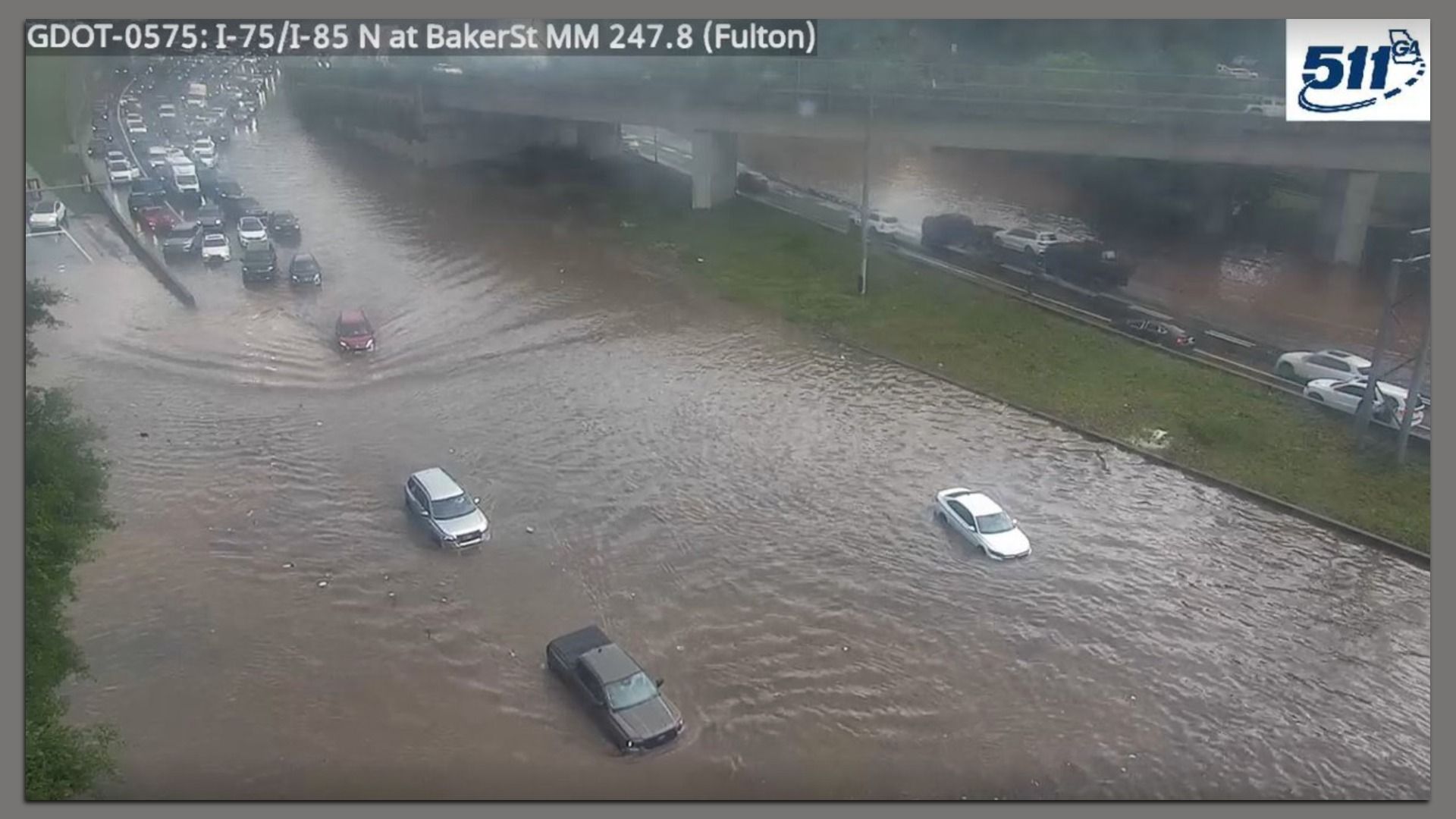 A surveillance camera image of a flooded interstate with several cars submerged. A long line of cars is backed up before the flooding begins and stuck. A green median separating lanes is visible.