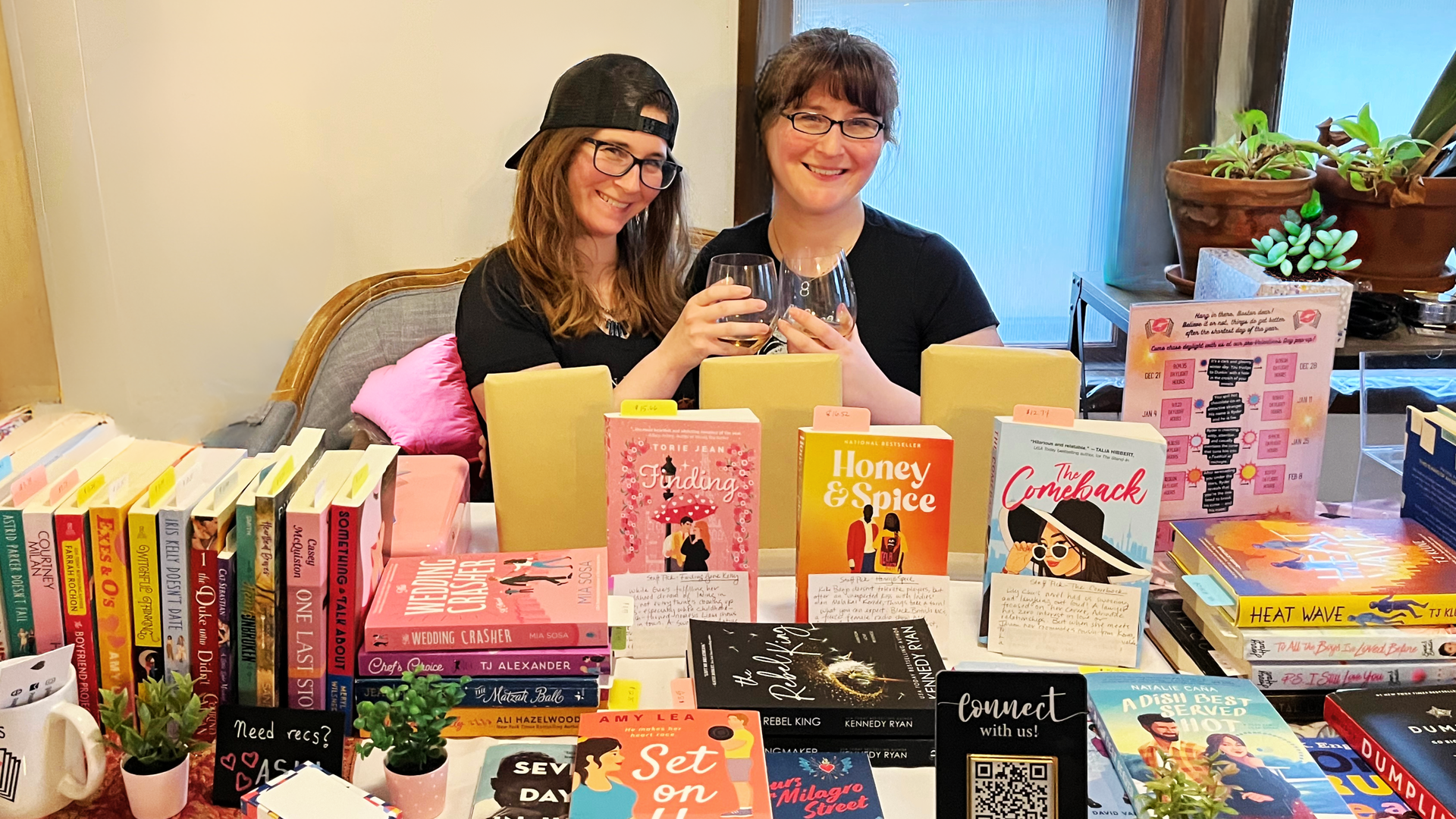 Twins Hannah, left, and Lily, right, Barrett raise winge glasses as they sit in front of a table filled with various romance books. They're starting a romance-themed book pop-up.