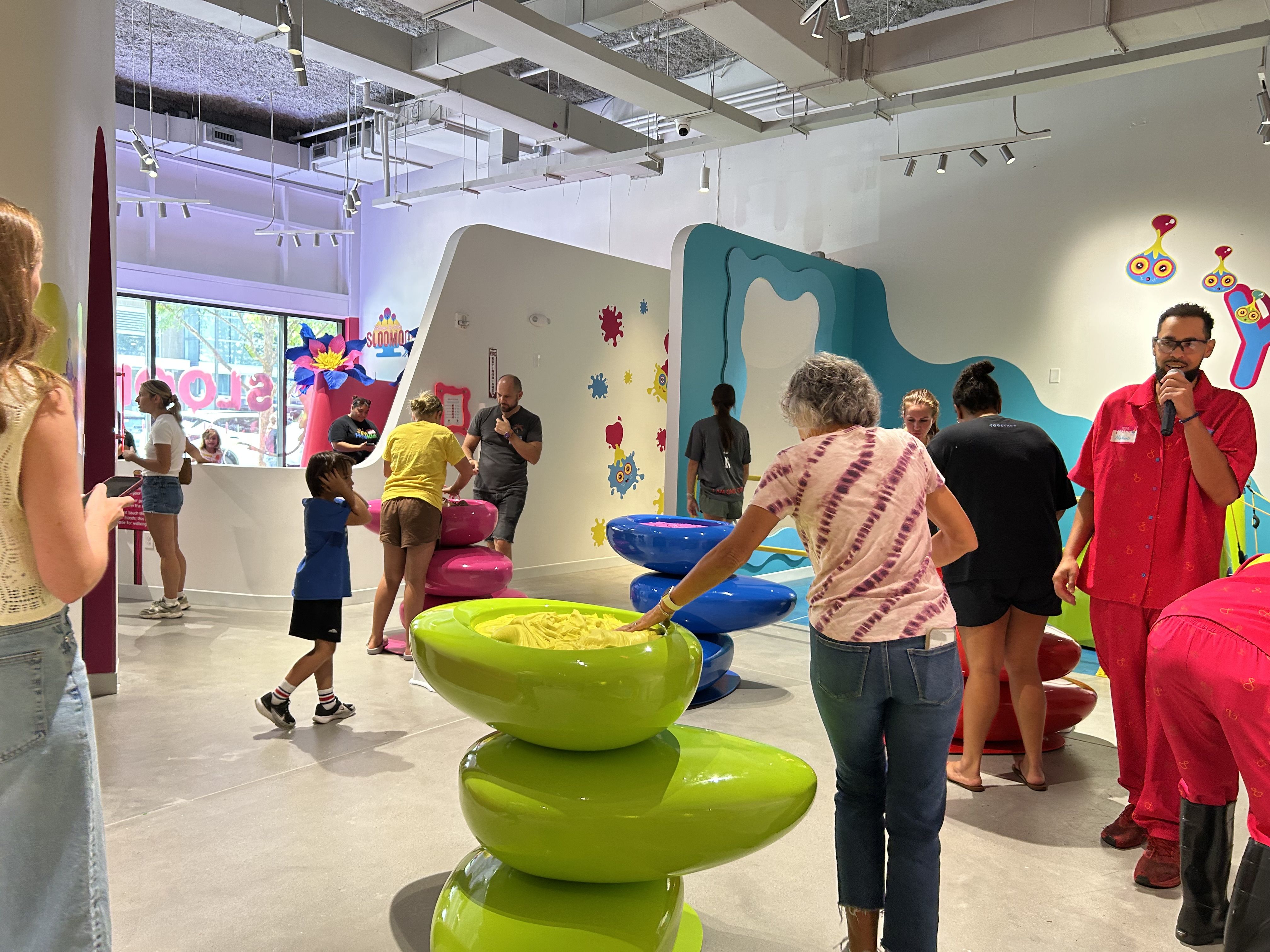 Bright indoor kids' play area with colorful curved walls and turquoise wave panels. Adults and children mingle; lime-green bowls stack in the foreground; a man in red speaks into a microphone.