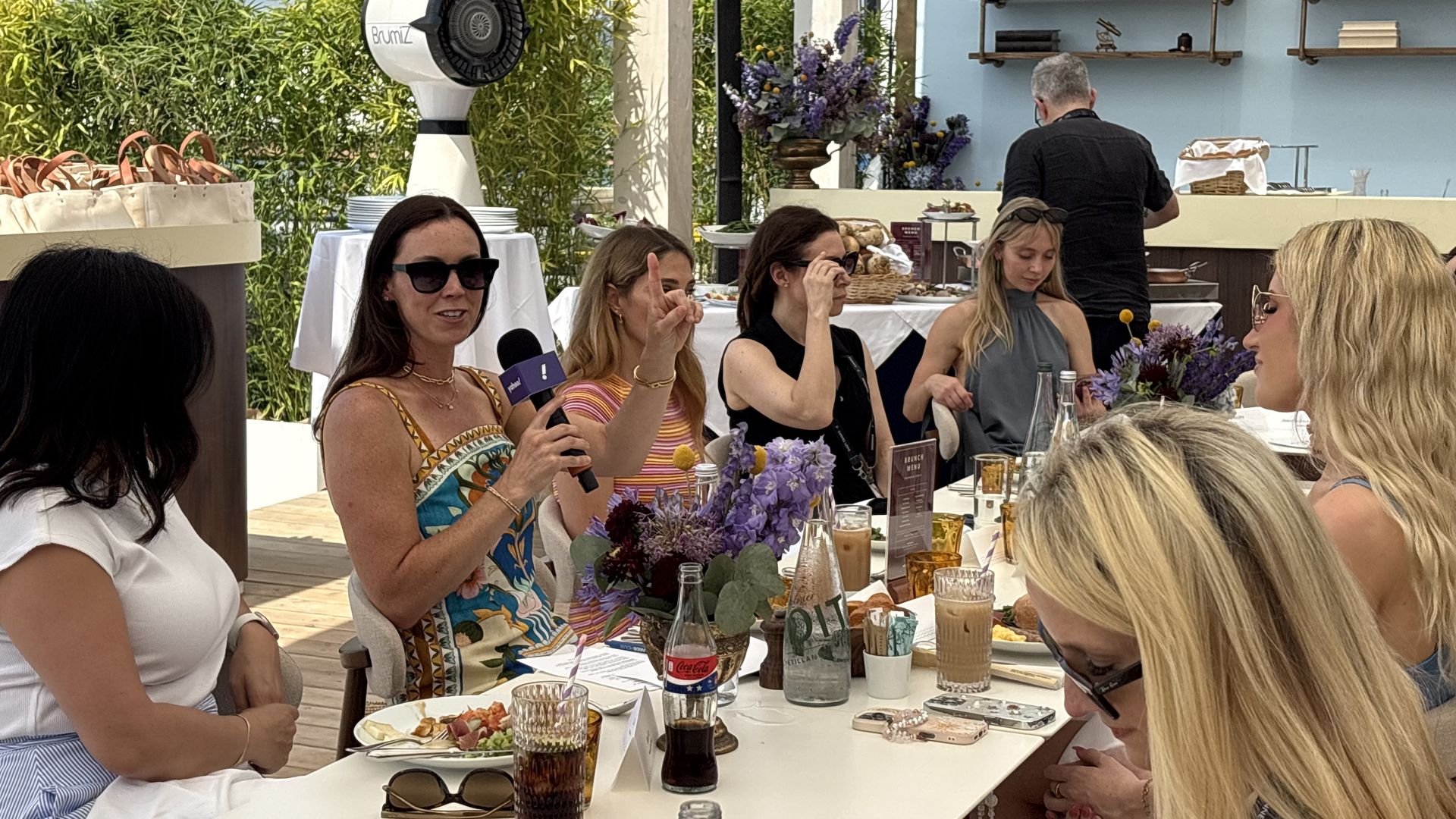 Group of guests dining outdoors at a white table with purple flowers; a woman holds a purple microphone while others chat, as a server works at a buffet against a blue wall.