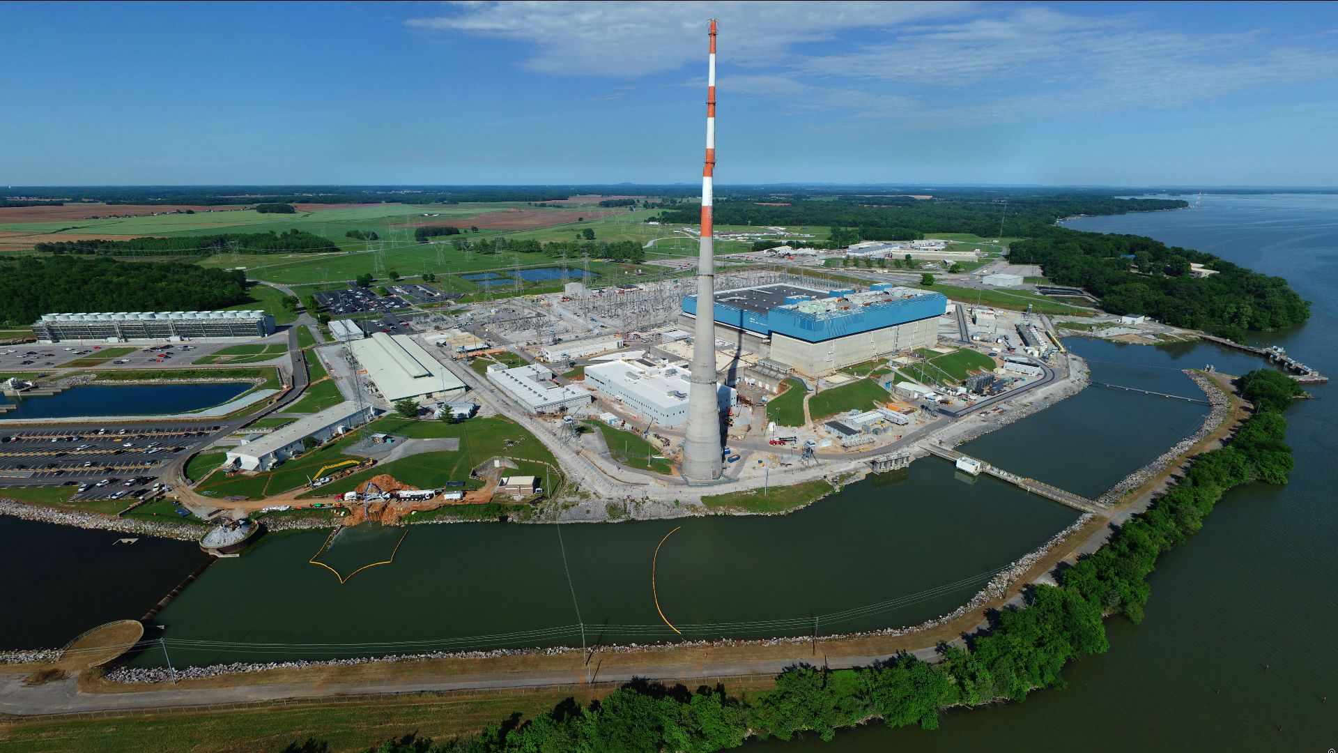 A large industrial plant with a tall red and white smokestack, surrounded by water channels and greenery under a clear blue sky.