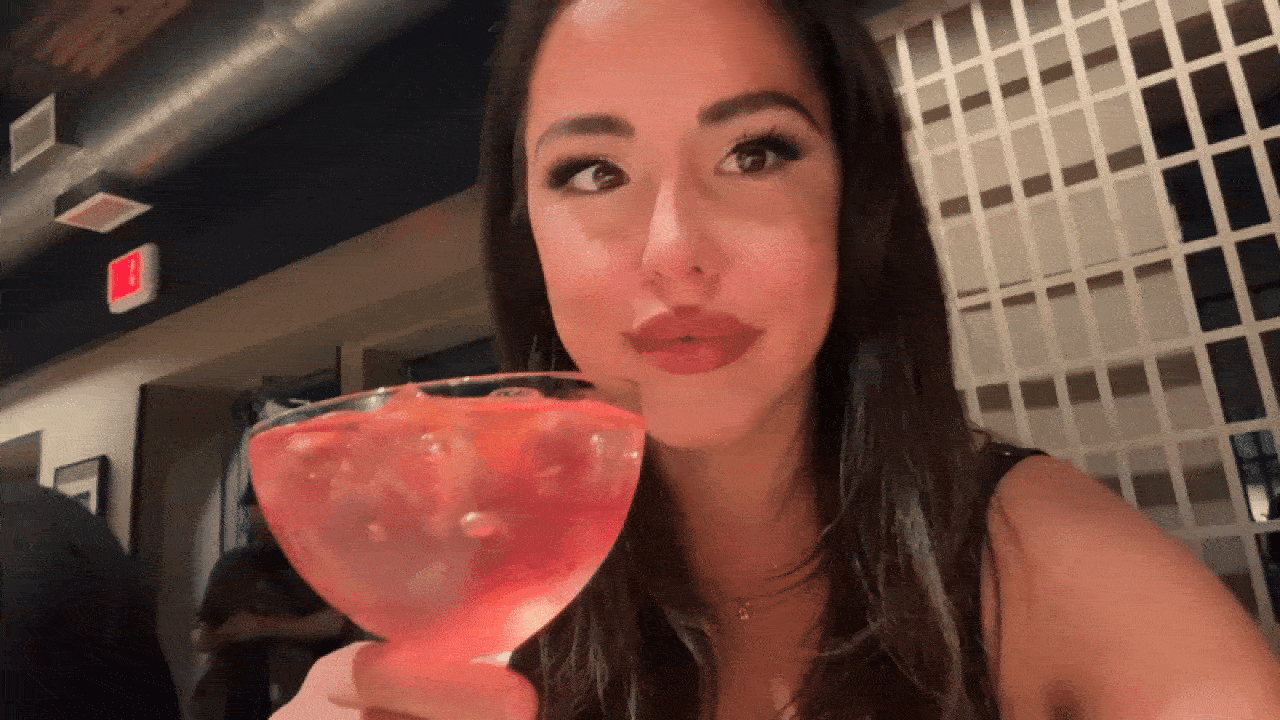 Woman with long dark hair holding a large cocktail glass filled with a pink drink and ice, sitting in a dimly lit restaurant with a lattice wall behind her.