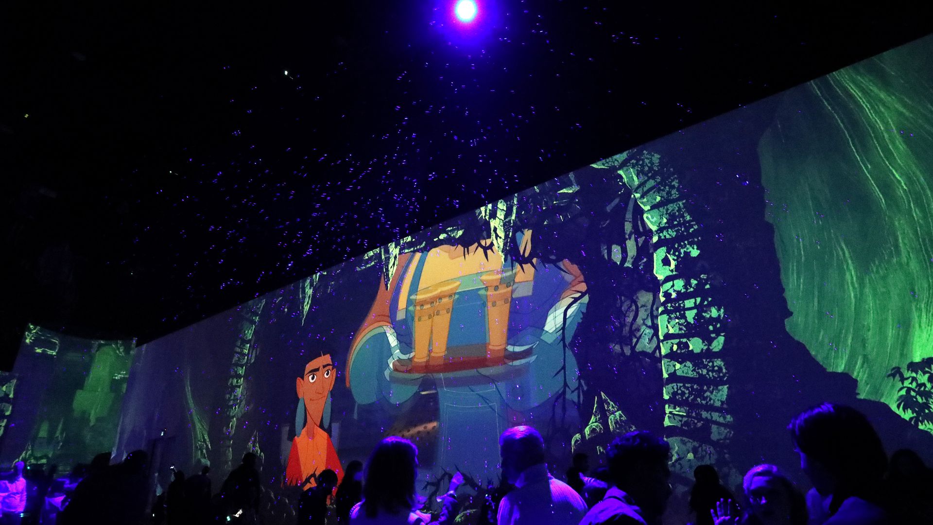 A small crowd of people watch floor-to-ceiling viewing of the "Emperor's New Groove" in a room dimly lit by a purple ceiling light.