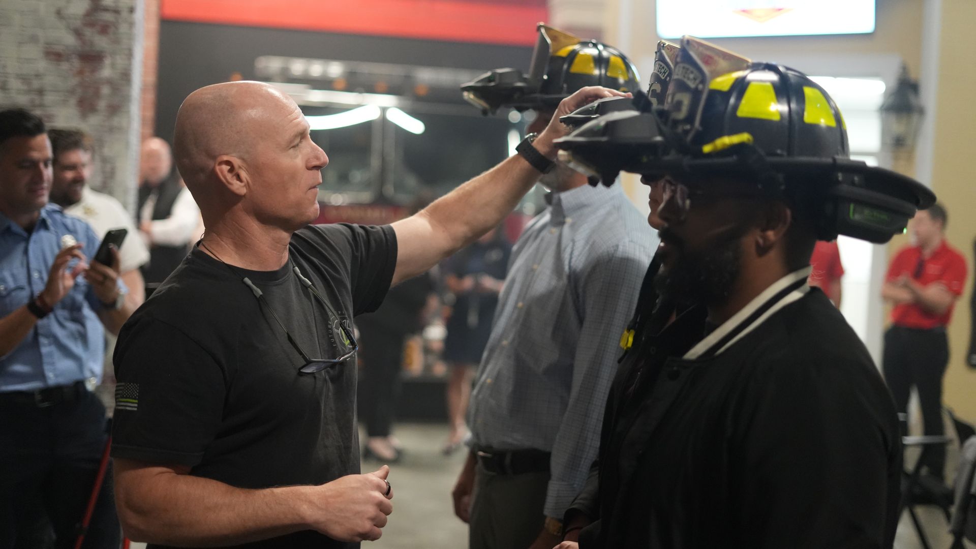 Indoor scene with a bald man in a black t-shirt presenting a firefighter helmet toward a row of yellow-and-black helmets on gear, as people watch in the background.
