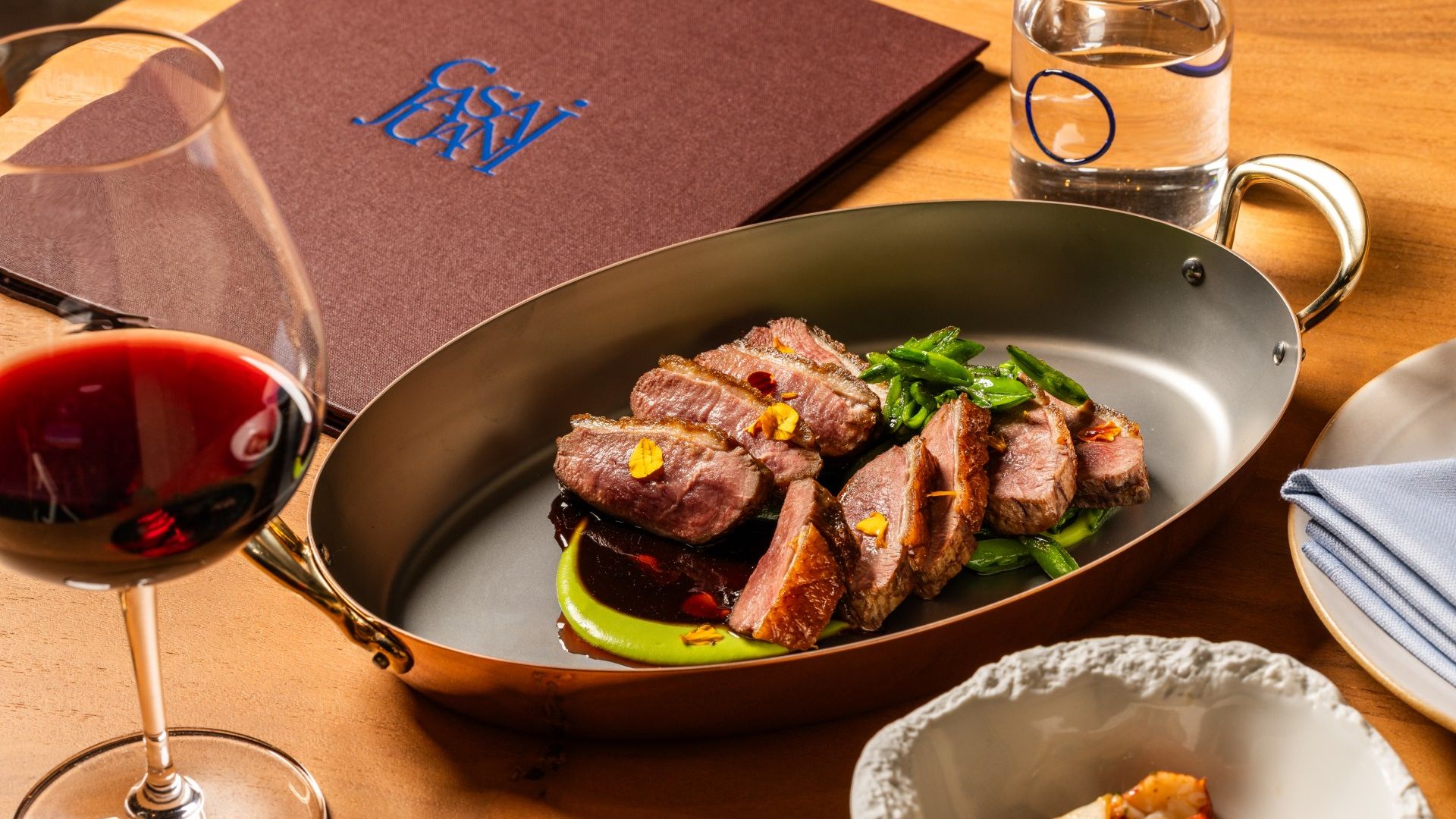 Pan with sliced duck on a green puree, topped with edible flowers and sauce, set on a wooden table beside a glass of red wine and a water glass; in background a menu with blue logo.