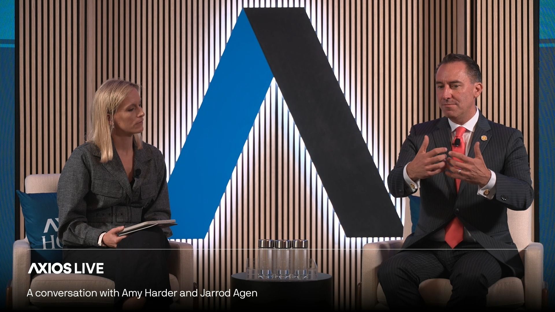 Two interviewers sit on a studio set with vertical wooden slats; left: blonde woman in a grey blazer with a notebook, right: man in a pinstripe suit gesturing. "AXIOS LIVE" logo.