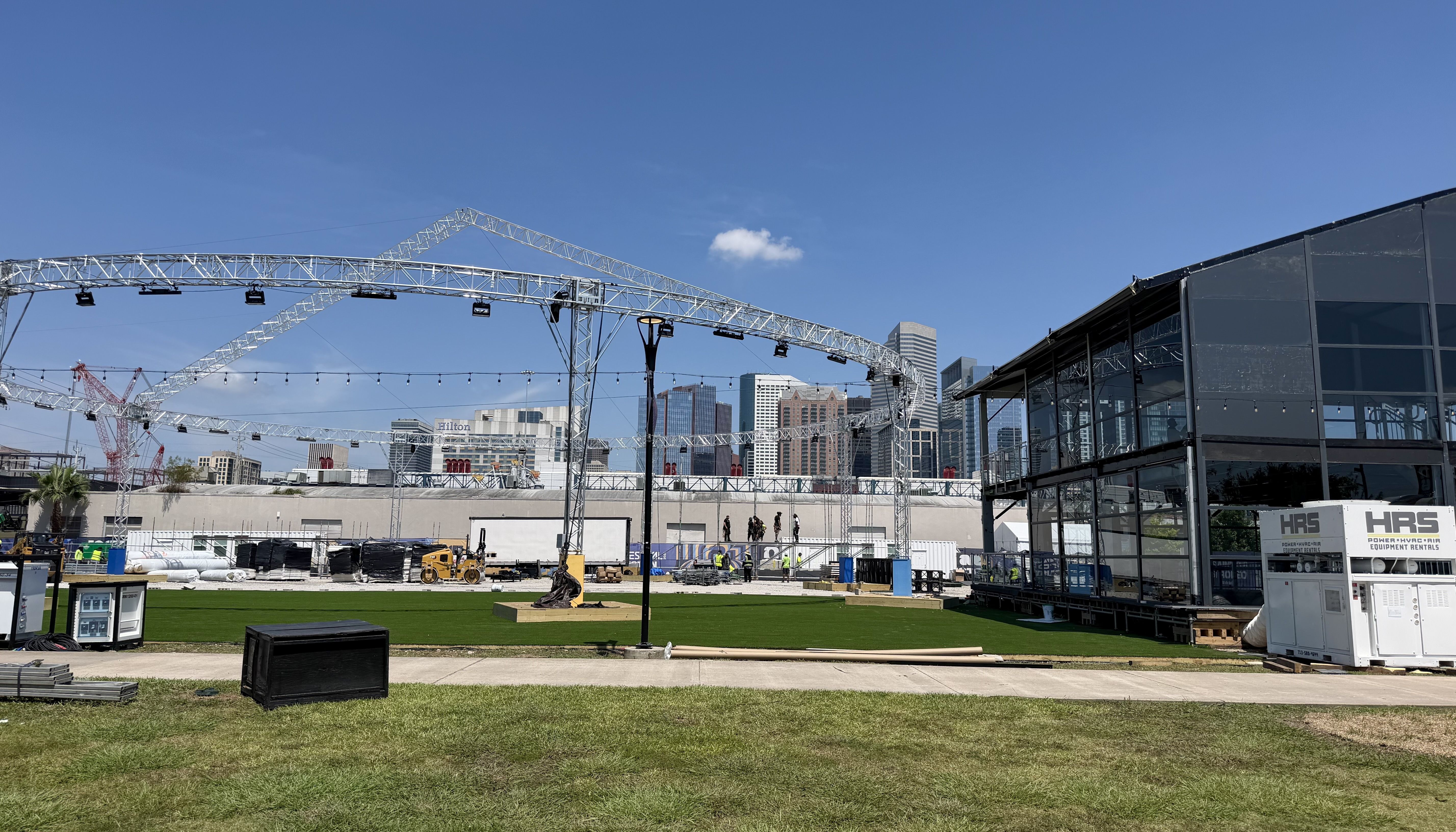 Construction site with a network of silver metal truss arches and hanging lights, a glass-walled building on the right, and a distant city skyline under a clear blue sky.