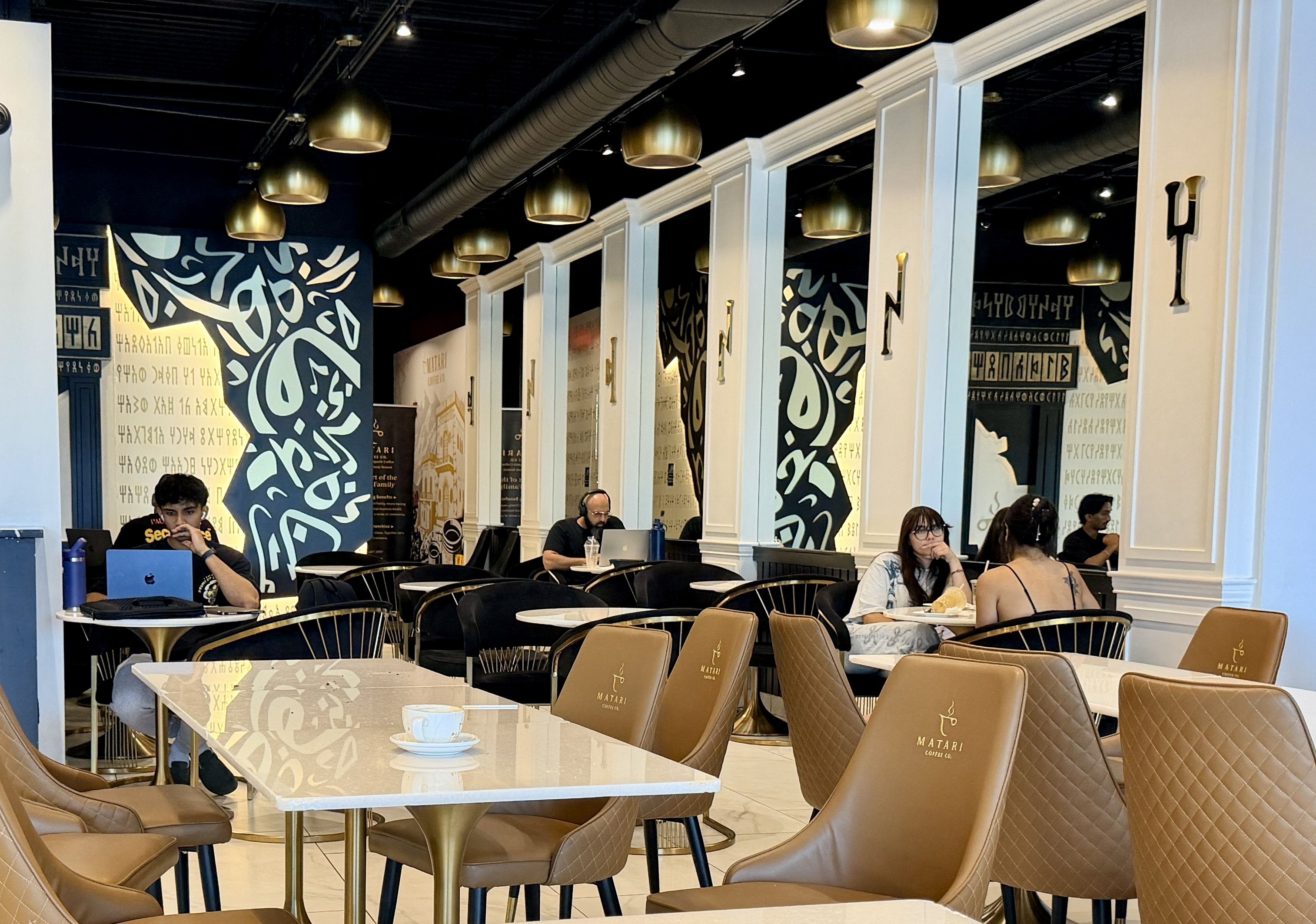Bright cafe interior with white divider walls, gold pendant lights, and blue patterned wall panels. People sit at marble tables on brown quilted chairs, working on laptops with coffee cups nearby.