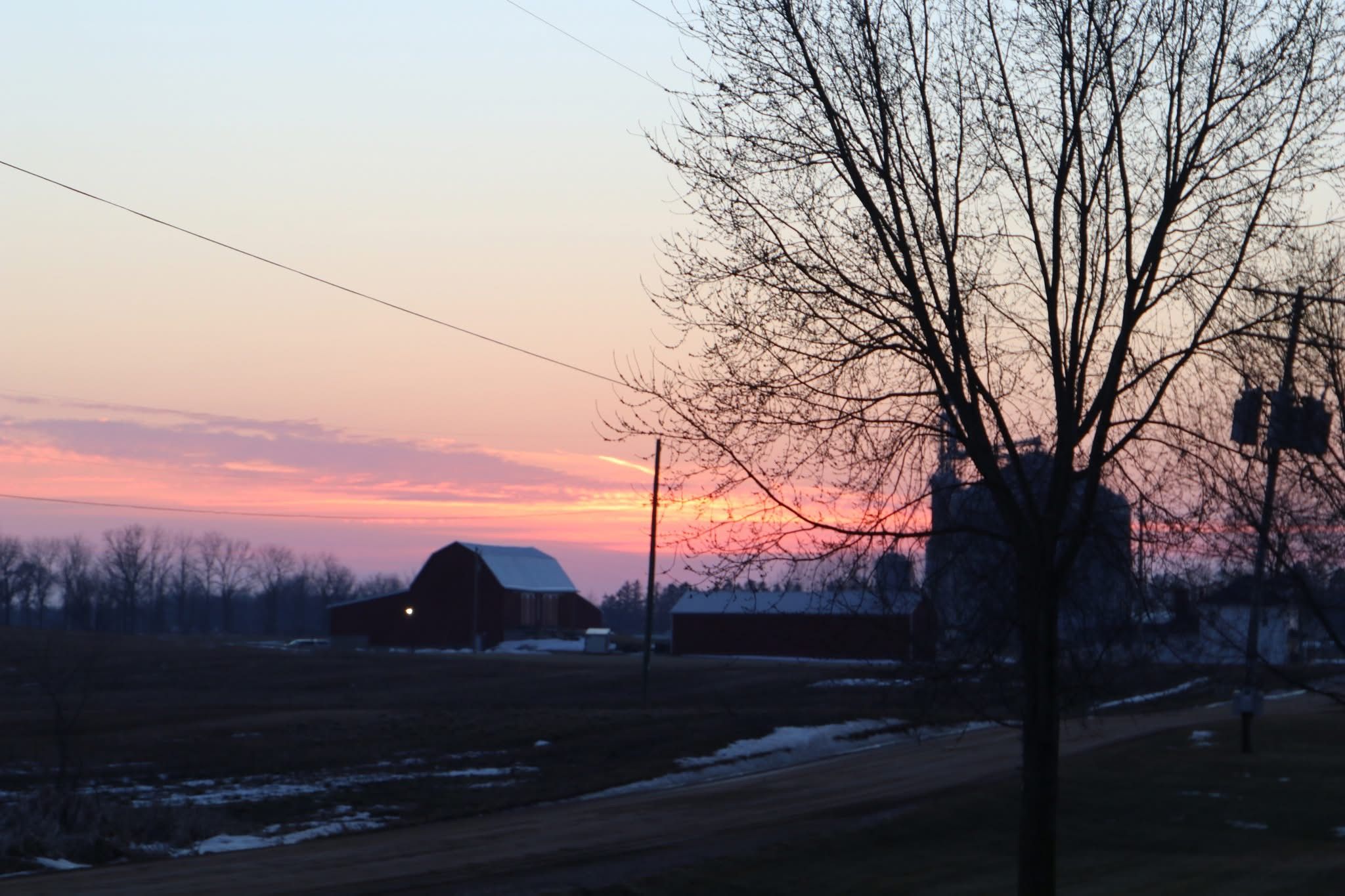 Rural scene at sunset with pink and orange sky, bare tree in foreground, red barns with some snow patches on ground, and a dirt road winding through the area.
