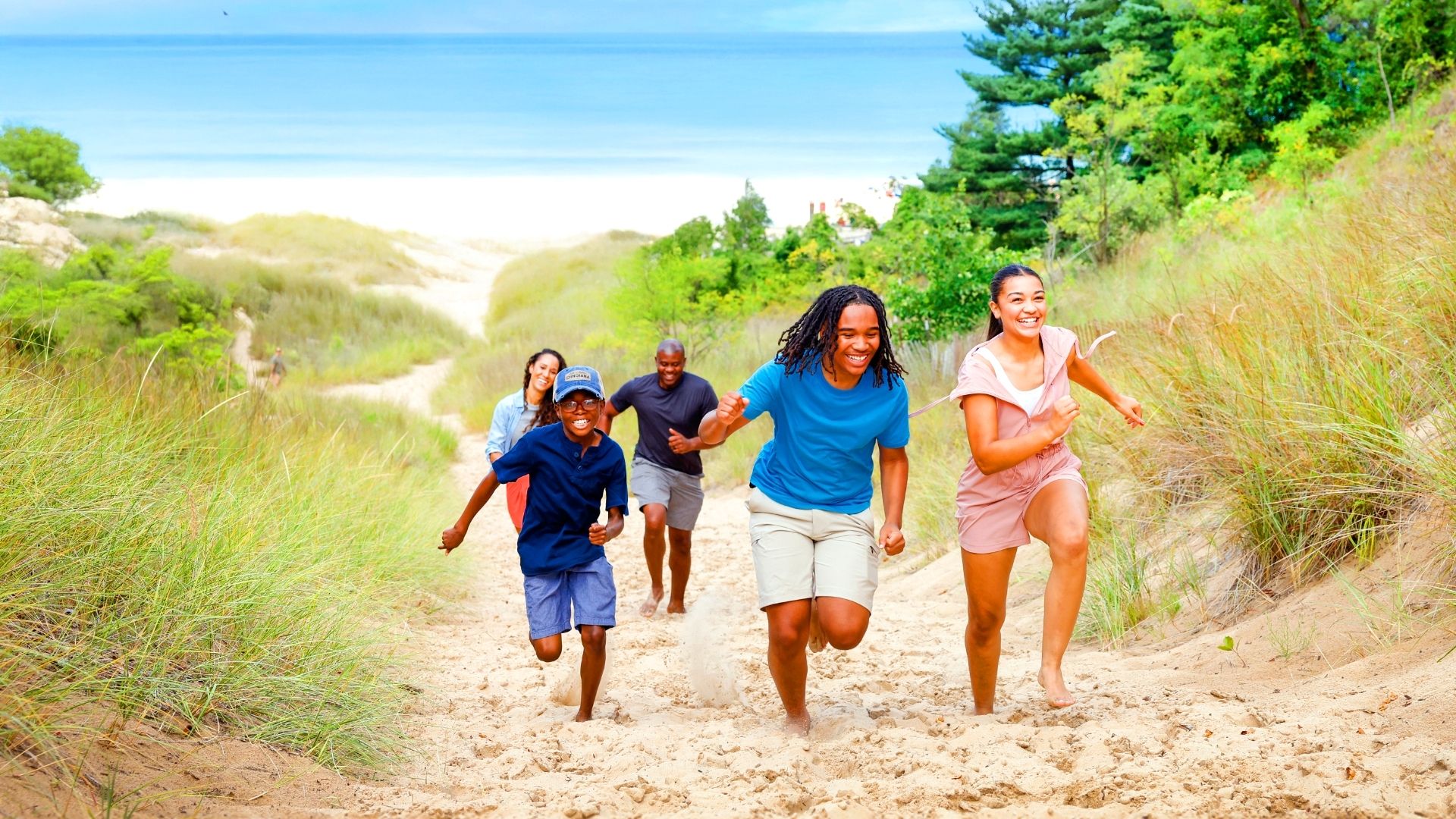 A smiling family joyfully runs uphill through sand dunes, with lush greenery and a serene lakeshore in the background.