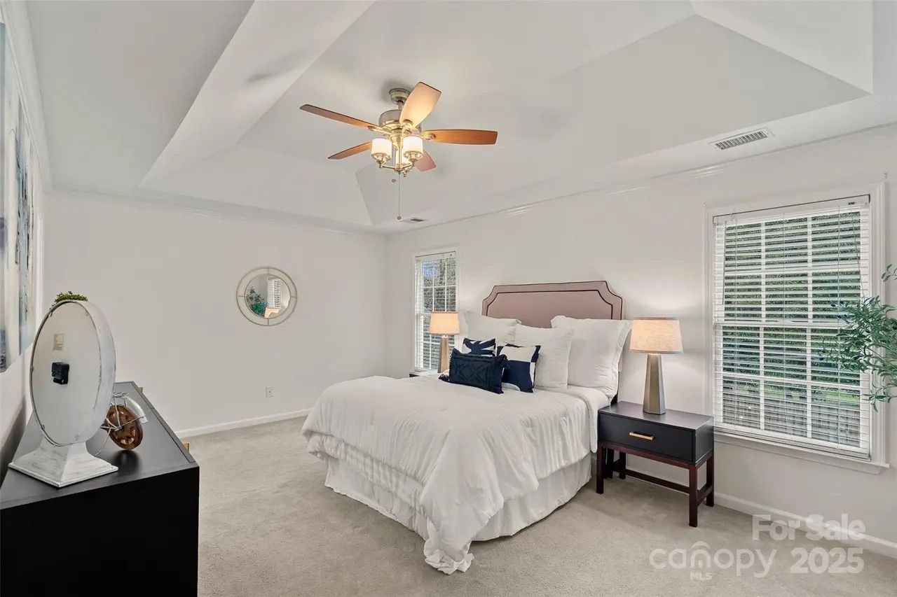 Bright bedroom with white walls, beige carpet, a bed with white bedding and navy pillows, dark wood nightstand with lamp, ceiling fan with lights, large windows, and decorative mirror.