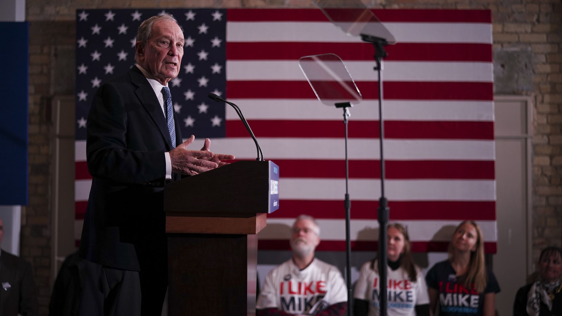 In this image, Mike Bloomberg stands in front of an American flag while on the 2020 campaign trail