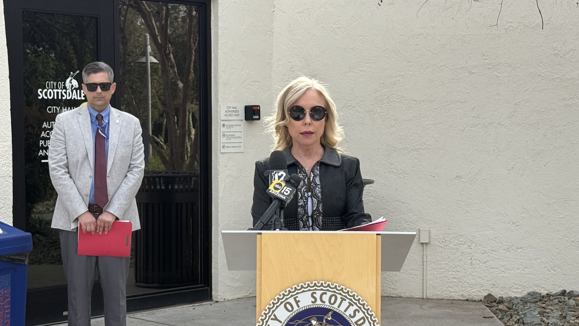 Woman in sunglasses and black jacket speaking at a podium with City of Scottsdale seal, alongside man in light gray blazer and maroon tie holding a red folder, outside city hall.