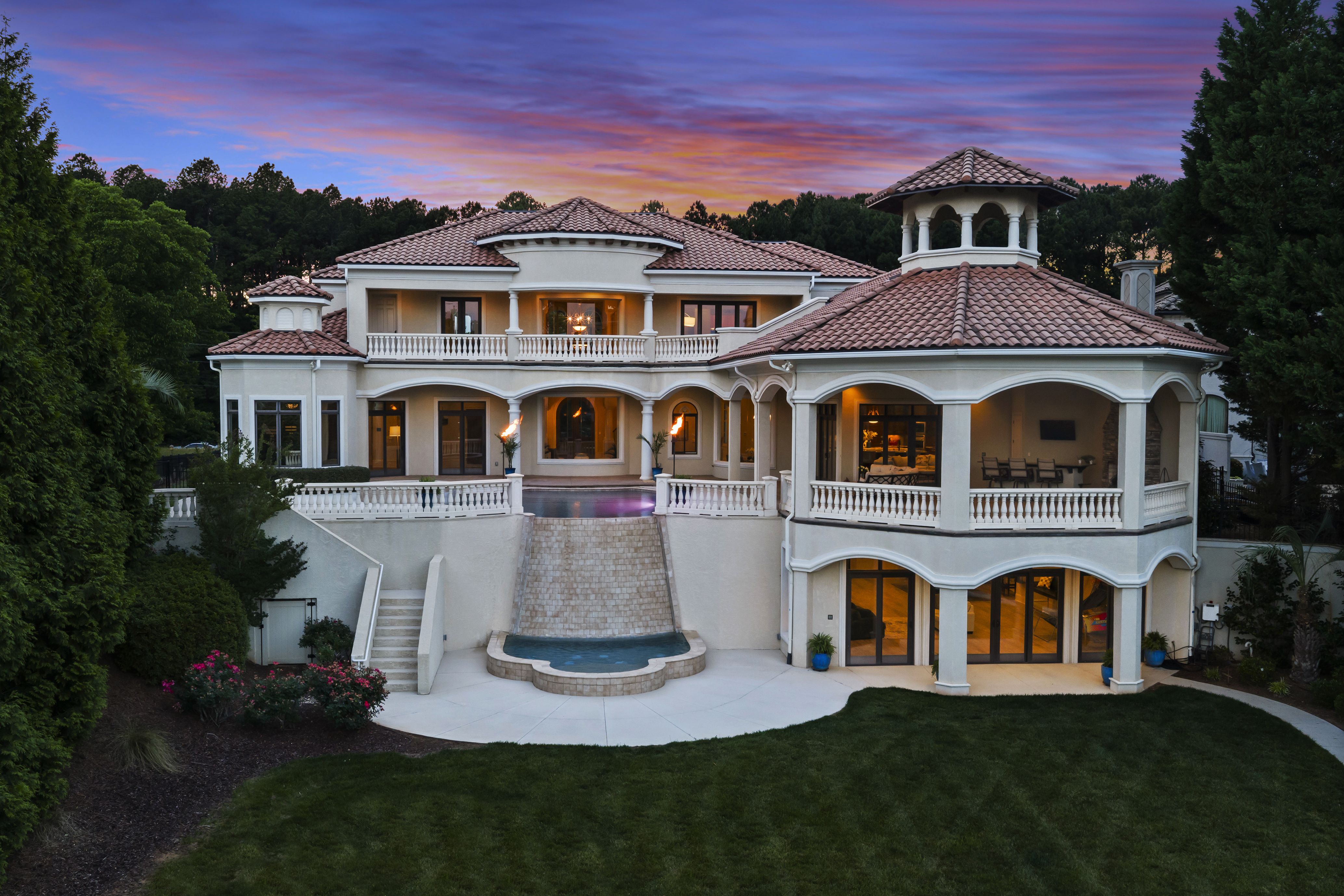 Grand Mediterranean villa with terracotta tile roof, white stucco, arched colonnades and a tower. Surrounded by trees, a curved courtyard pool glows under a pink, orange, purple sunset.