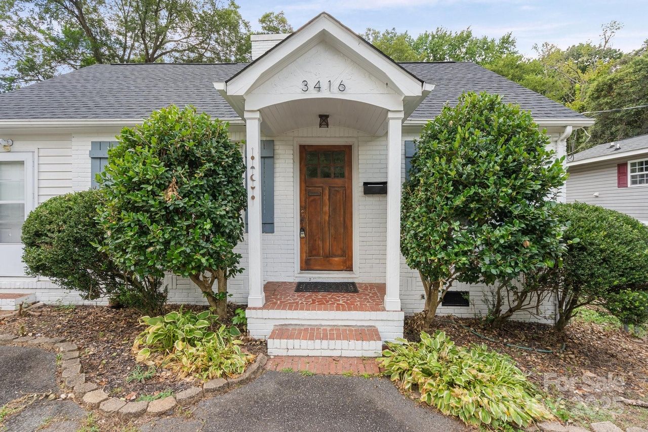 White brick house entrance with a wooden door, brick steps, two large green bushes on either side, house number 3416 above porch, and small plants near the walkway.