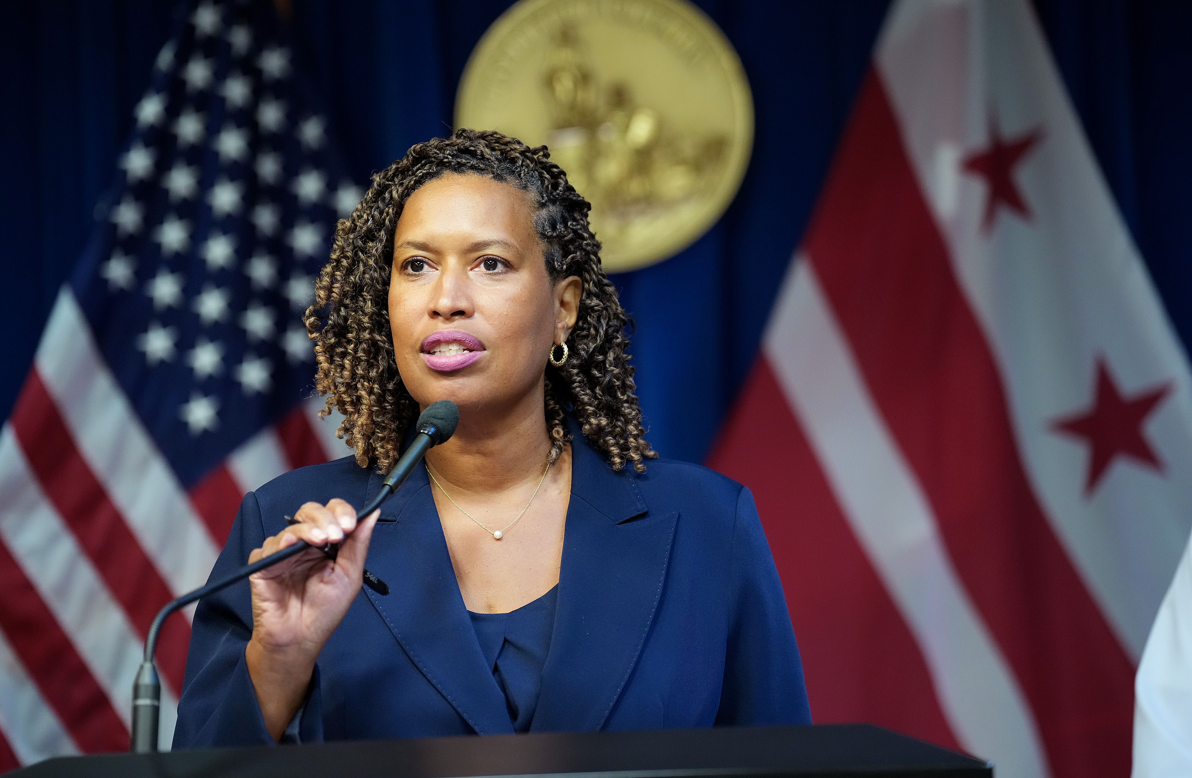 Woman with curly hair wearing a navy blue suit speaking at a podium with a microphone, with American and Washington, D.C. flags in the background.