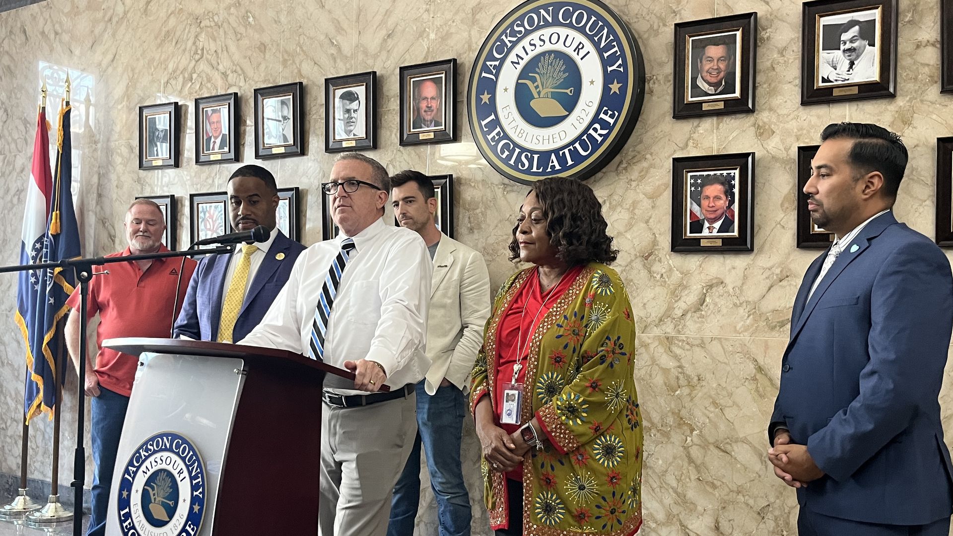 A group of six diverse officials stand at a podium with a microphone in front of Jackson County Legislature seal and portraits on a marble wall with Missouri flags to the left.