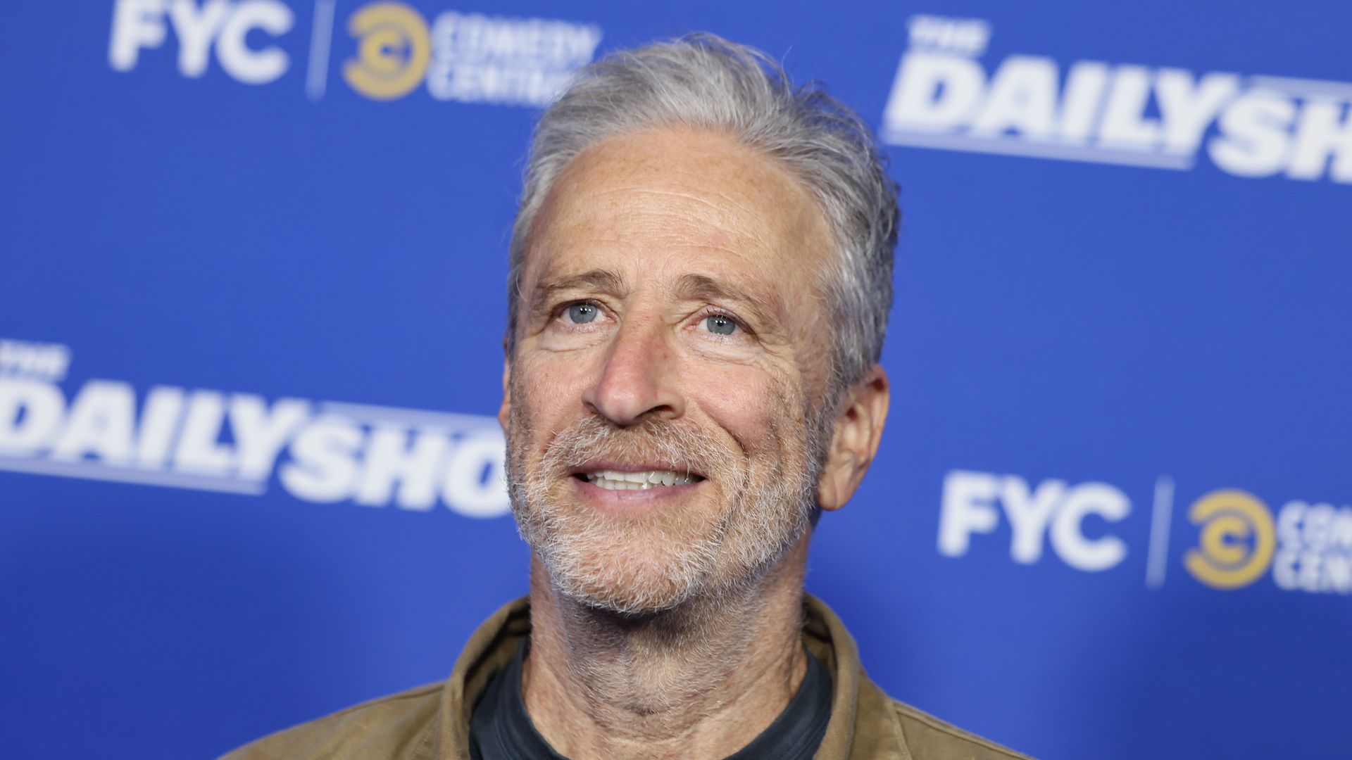 A gray-haired Jon Stewart, wearing a brown, suede jacket and black t-shirt with stubble, smiles in front of a "Daily Show" sign in Los Angeles, California.