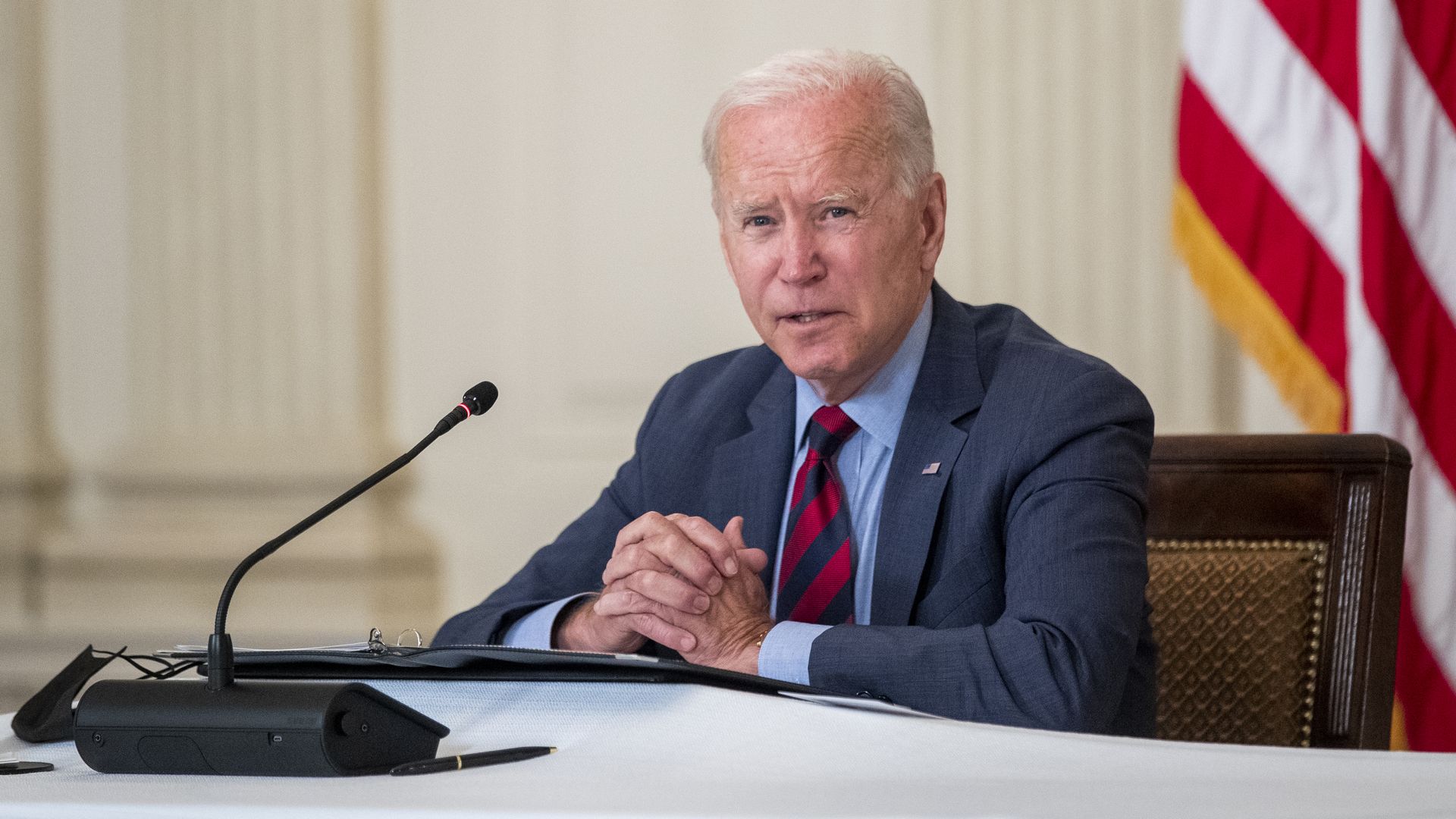 President Joe Biden sits at a microphone with this hands clasped together during a meeting. 