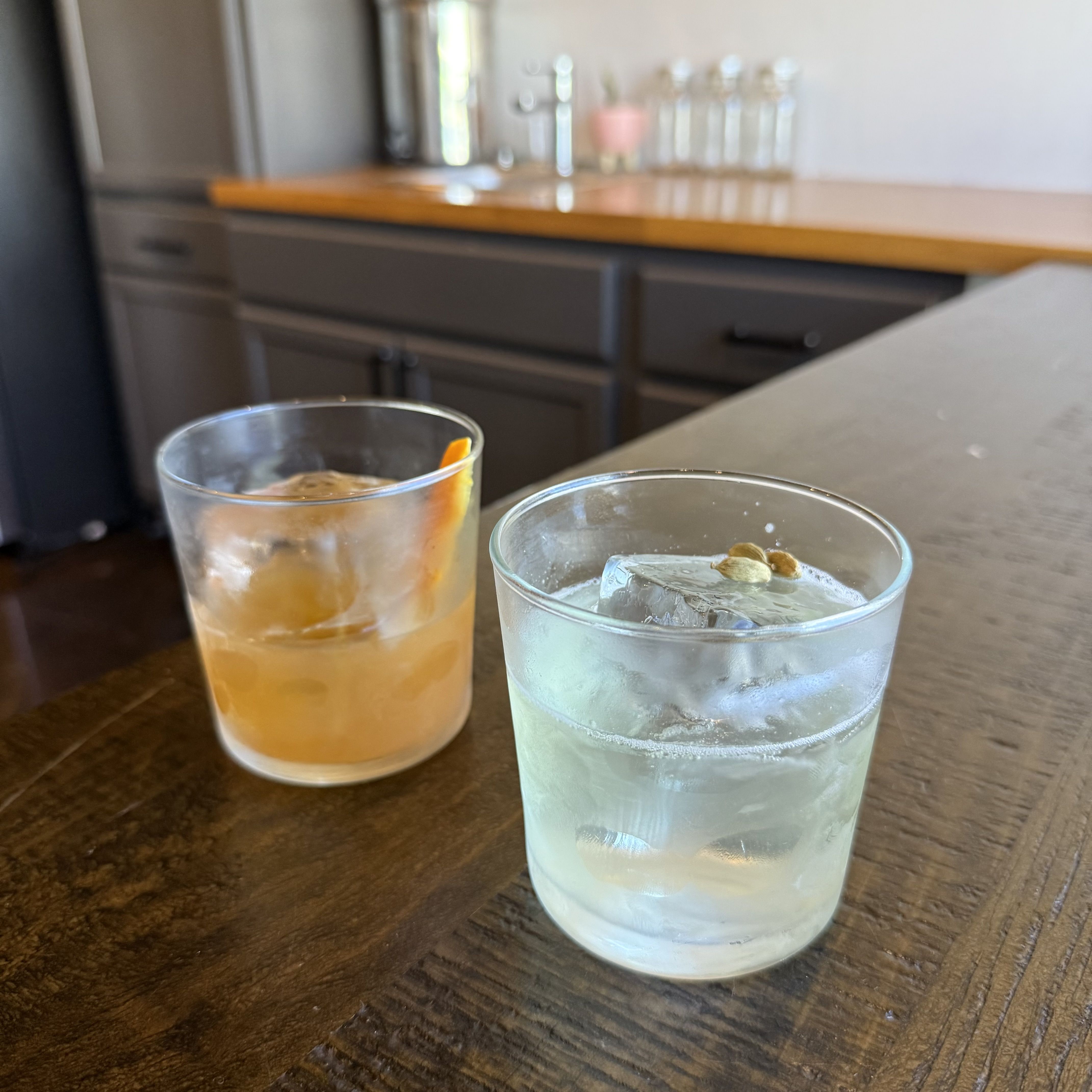 Two short tumblers on a dark wooden bar. Front glass has a pale blue drink with large ice; behind it is an amber drink with ice and an orange twist. A blurred kitchen sits in the background.