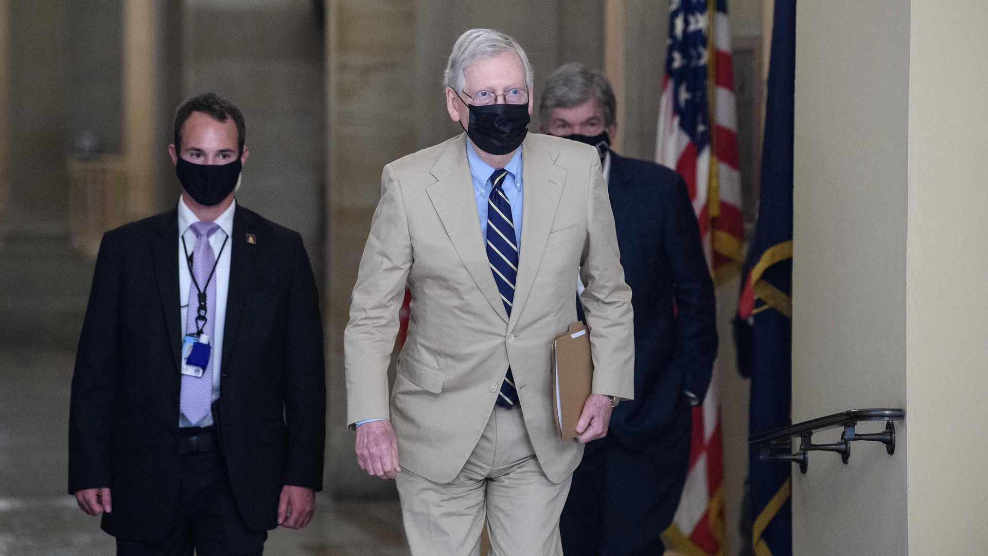 Mitch McConnell wears a tan suit as he walks through Congress with two men in black suits behind him