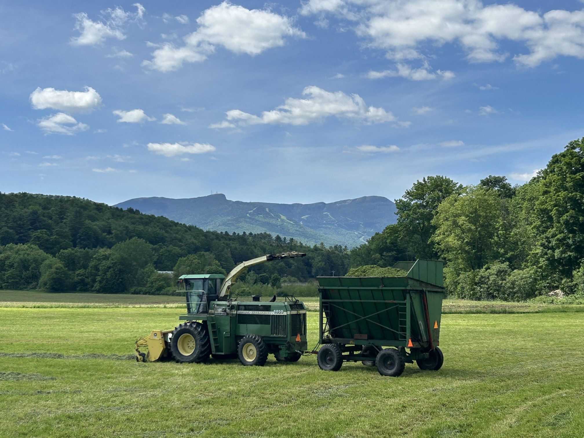 A green combine harvester and a large green grain trailer sit on a grassy field, with forested hills and distant mountains under a blue sky dotted with white clouds.