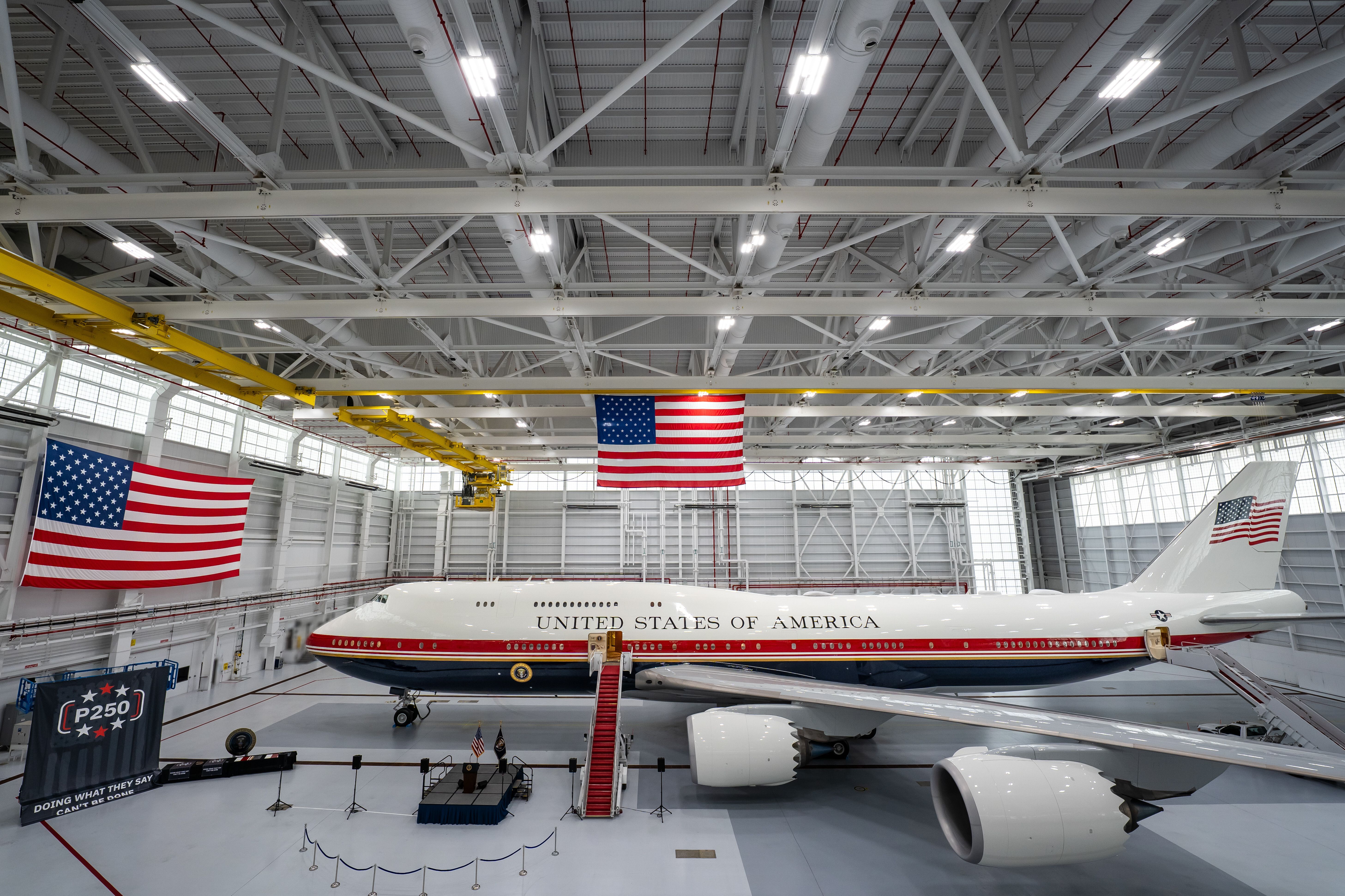 Inside a large hangar with white steel beams and bright lights, a United States airplane sits on the hangar floor, its fuselage marked UNITED STATES OF AMERICA, with American flags nearby.