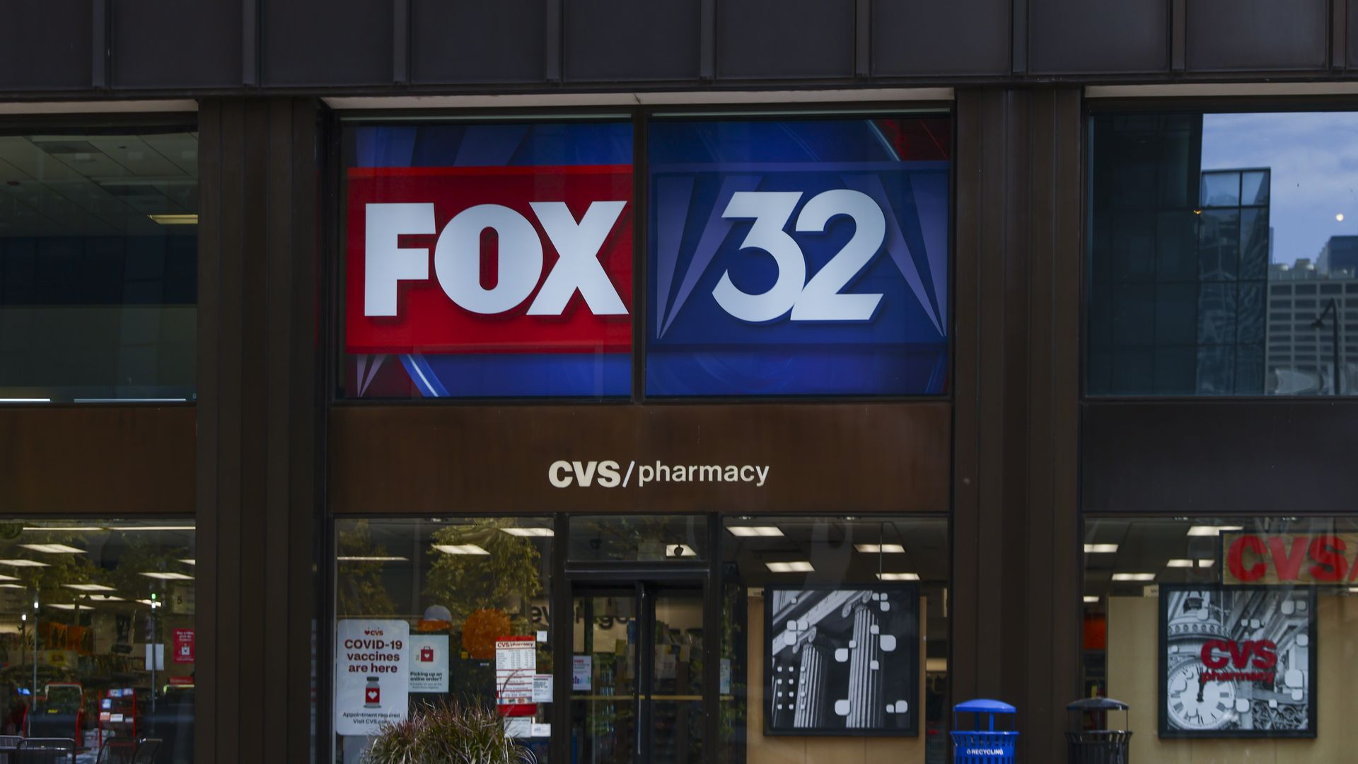 Storefront with large window sign reading "FOX 32" in red and blue panels; CVS/pharmacy logo beneath, glass facade, with flower planters in the foreground.