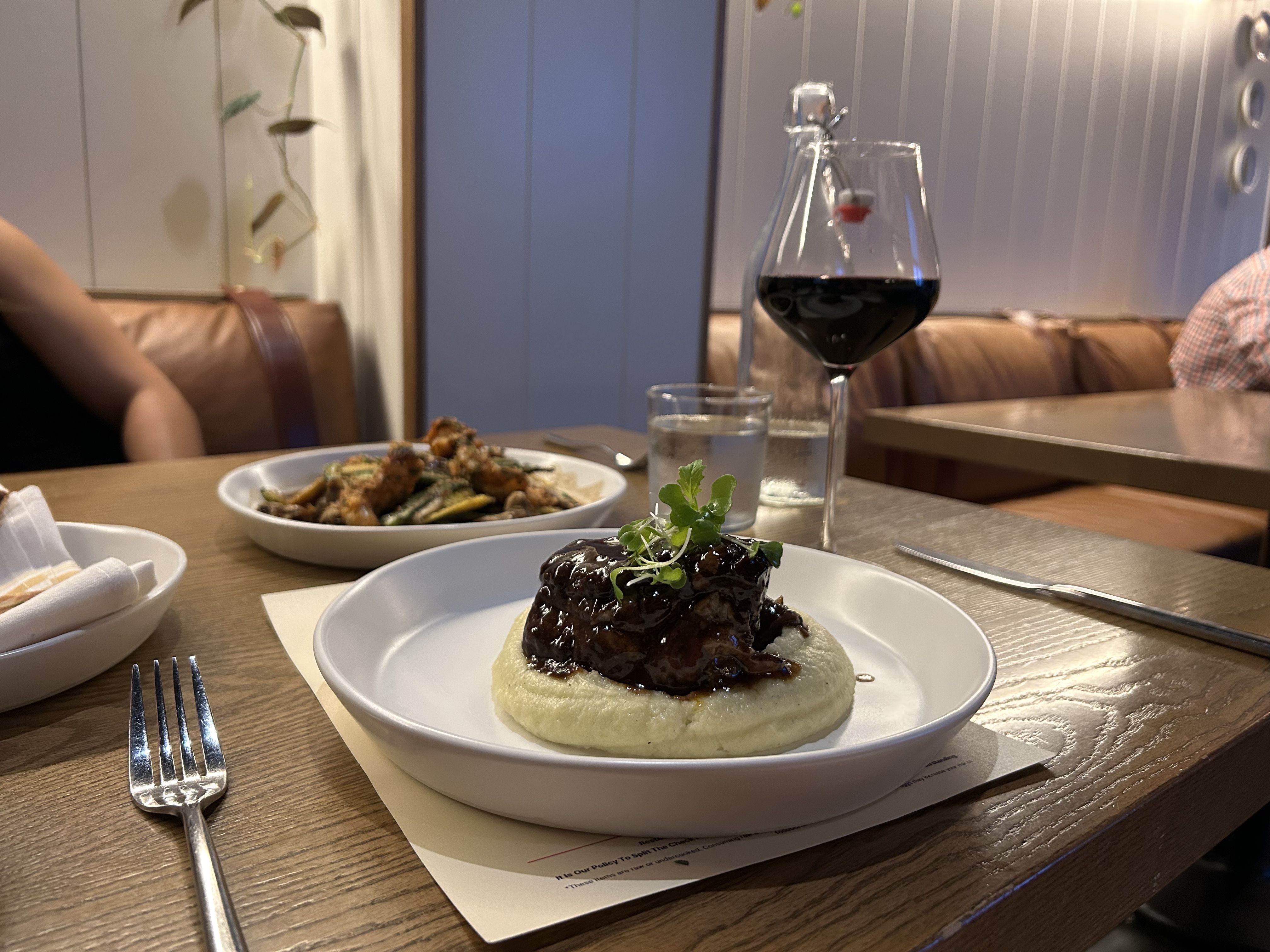 A plated dish featuring a piece of glazed meat on a bed of creamy mashed potatoes, garnished with fresh herbs. In the background, another dish with what appears to be fried or roasted vegetables. 