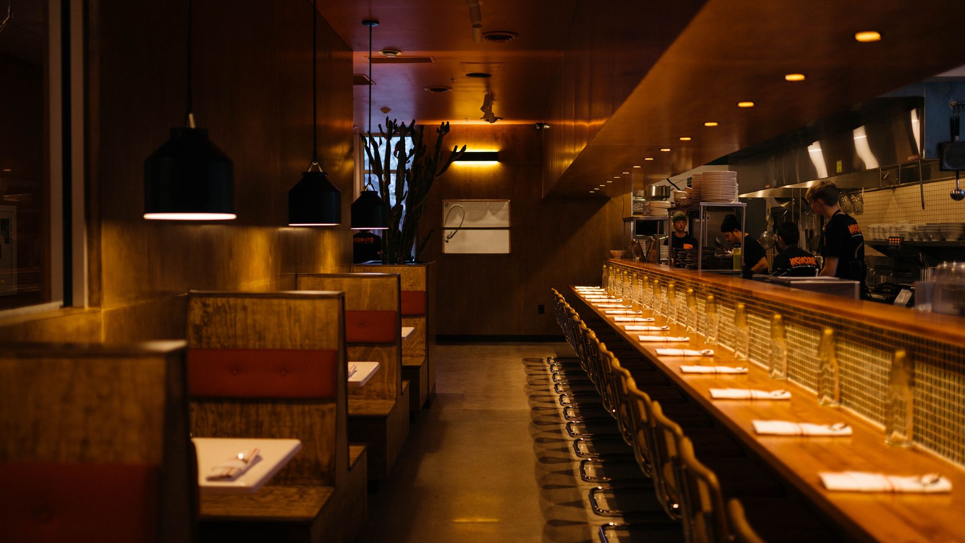 Dimly lit restaurant interior with a long wooden counter and row of stools on the right, warm pendant lights, booths on left, and a busy open kitchen with chefs in the background.
