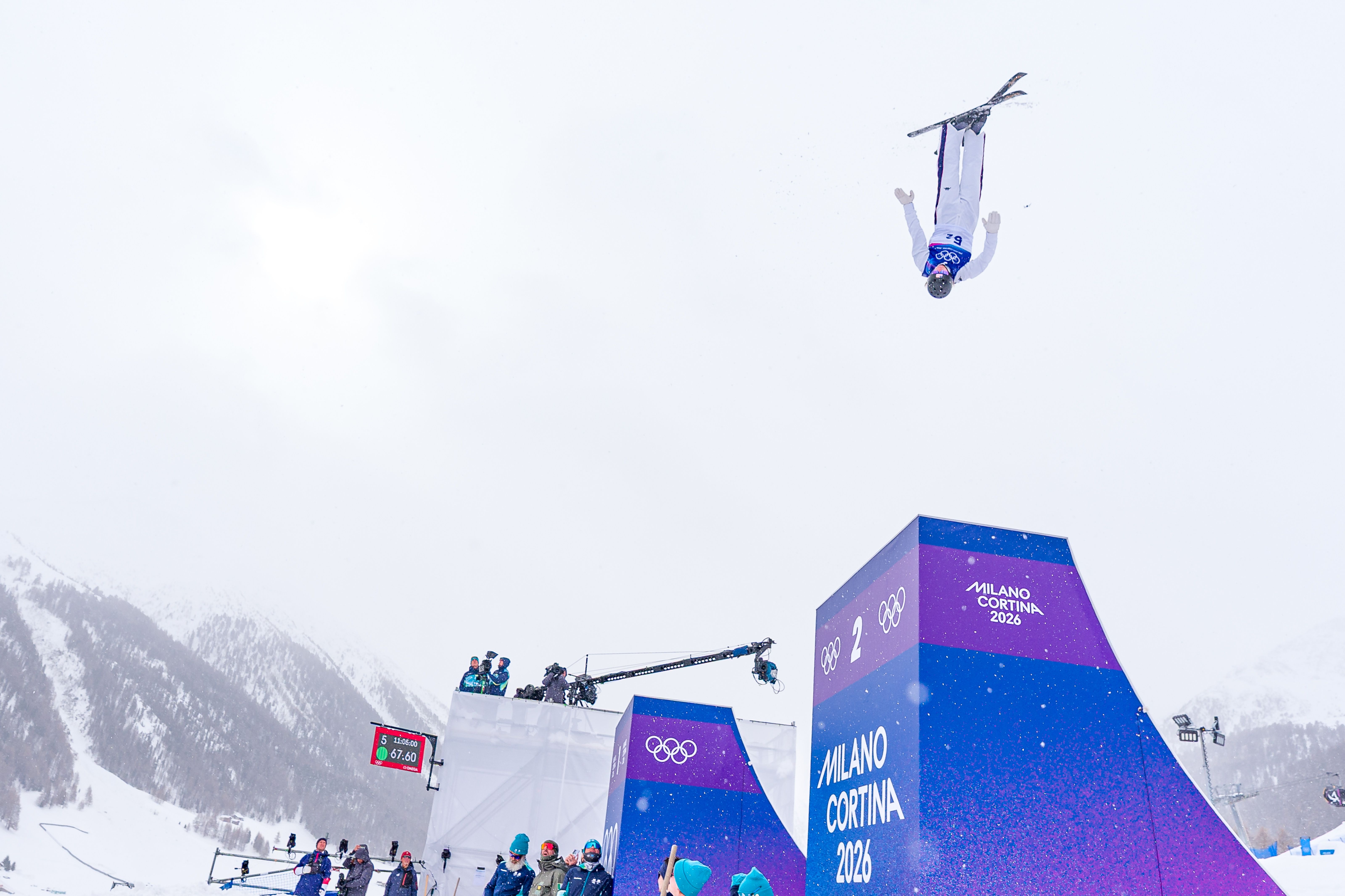 Skier Connor Curran in a white suit and helmet performing a mid-air flip above a purple ramp with "Milano Cortina 2026" and Olympic rings in snowy mountainous winter landscape.