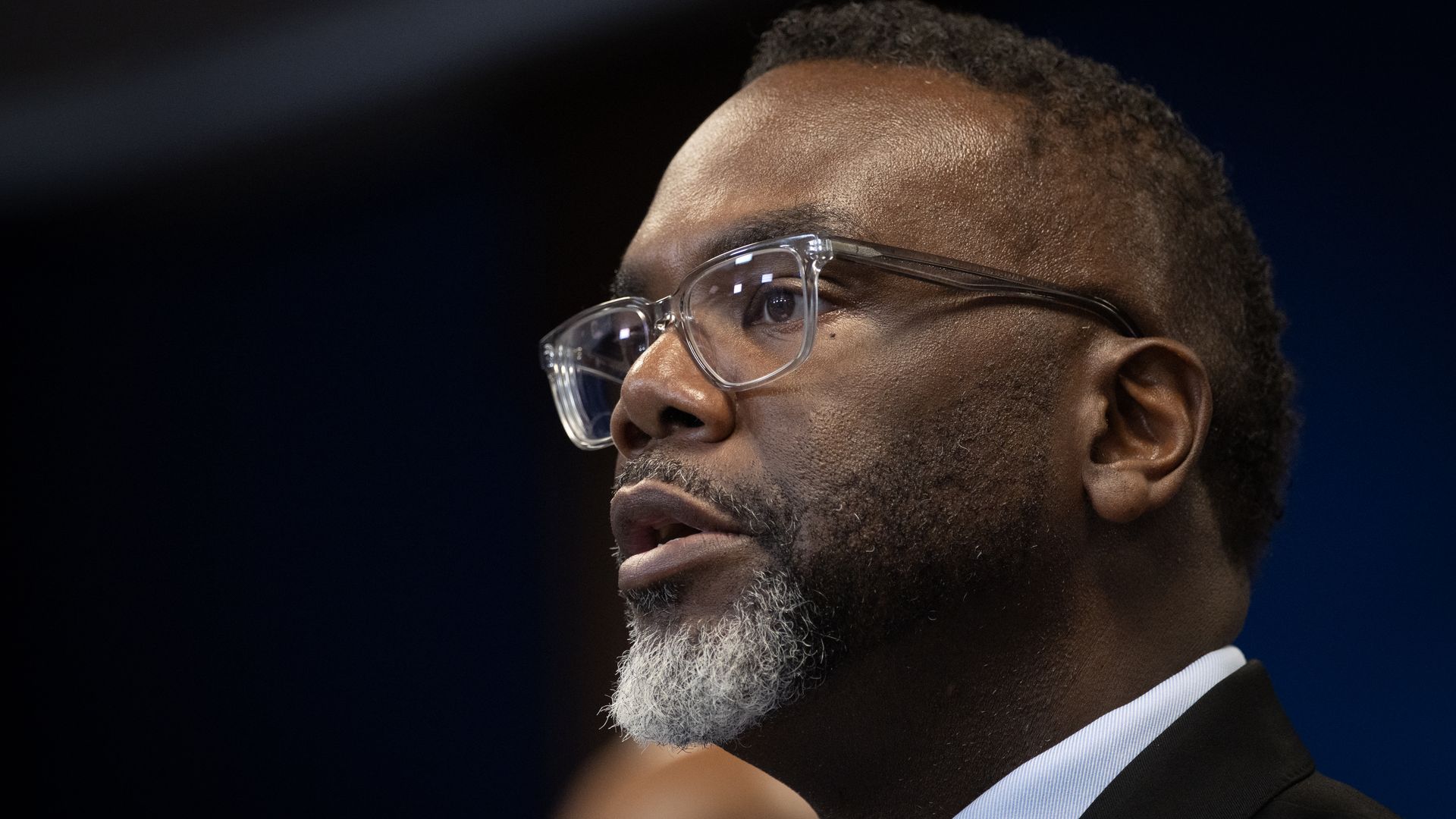 Close-up of a man with clear glasses, black and gray beard, and short hair, speaking with a focused expression, wearing a black suit and light blue shirt, against a dark background.