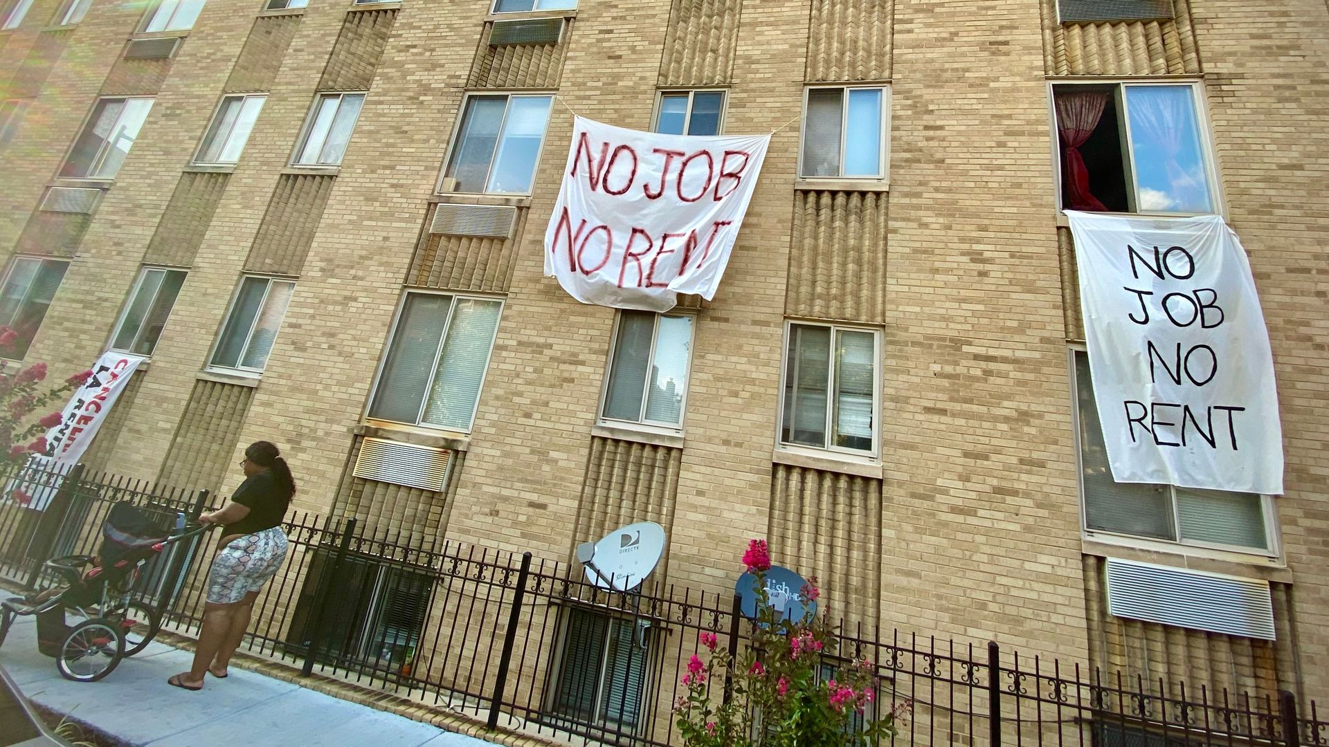 A woman and child stand in front of an apartment building. Signs hanging out the window assert 