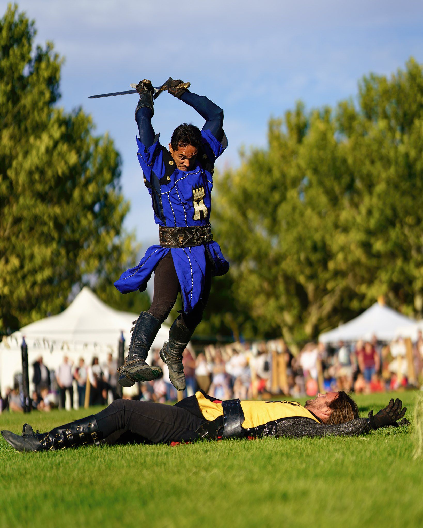 A man dressed as a medieval crusader leaps over another man while swinging a sword.