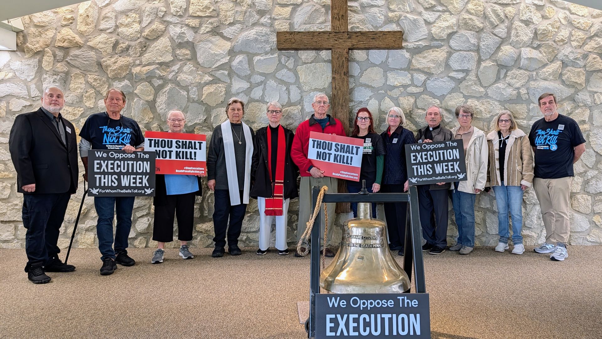 A group of thirteen people standing inside a chapel with a stone wall and wooden cross behind them, holding signs opposing execution and the death penalty, with a large bell in front.