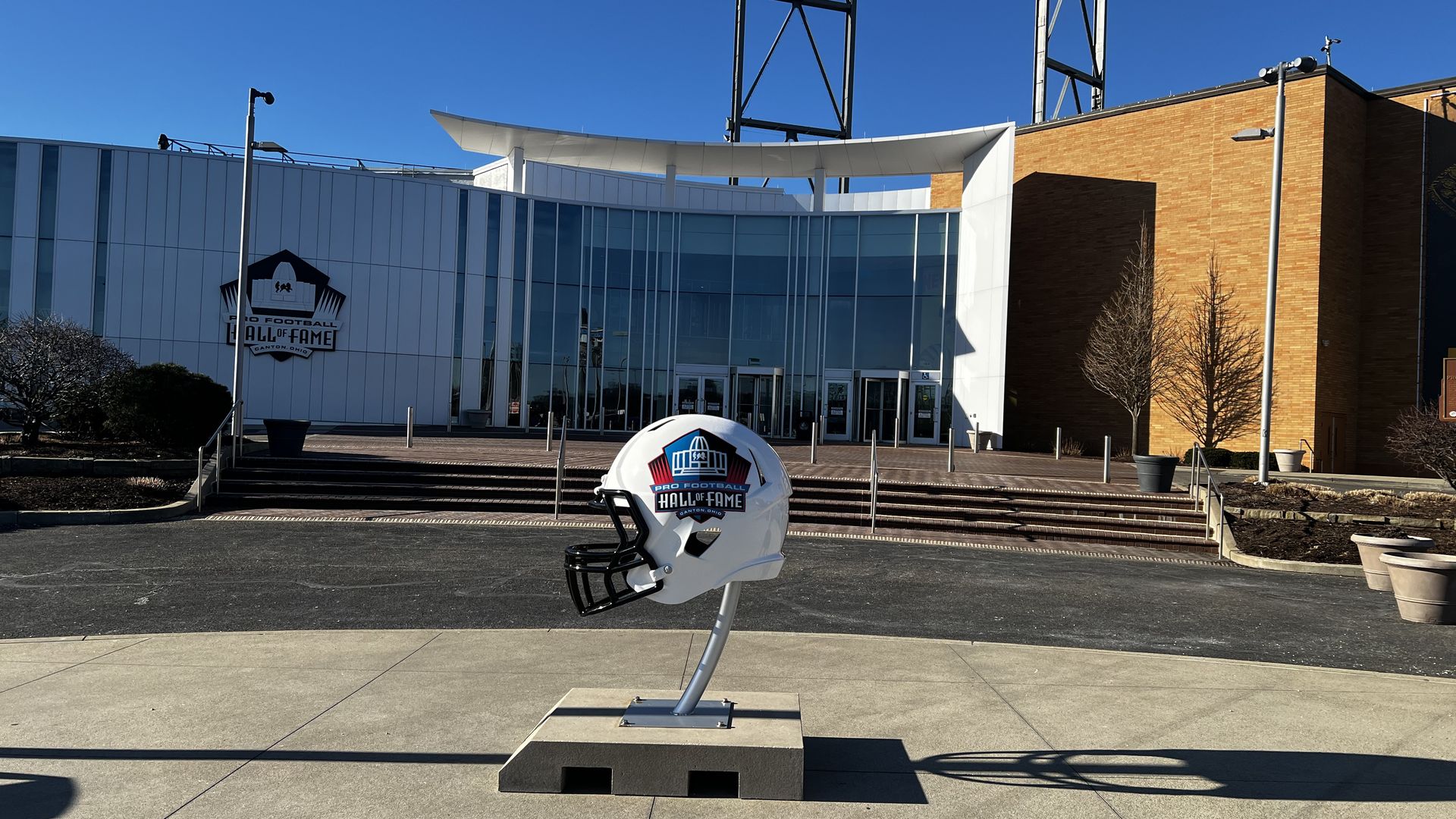 Front view of the Pro Football Hall of Fame in Canton, Ohio, featuring a curved white canopy over glass doors, a large football helmet display, brick wall on the right, and a clear blue sky.