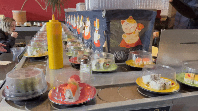 A moving image showing a sushi restaurant, with plates of items moving on a conveyor belt as a patron eats.