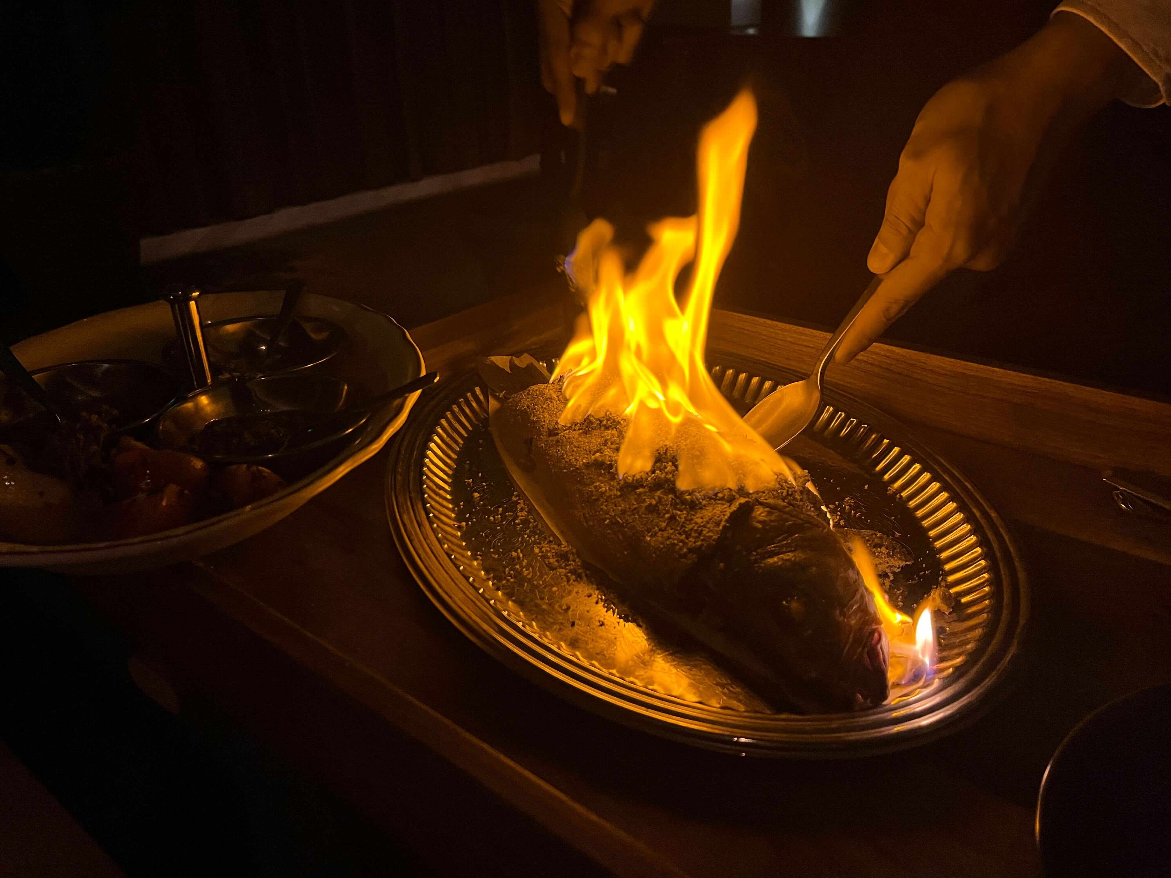 Whole fish covered in spices being flambéed on a silver platter with bright orange flames, hands holding utensils preparing it, and a bowl with sauces on the side in low light.