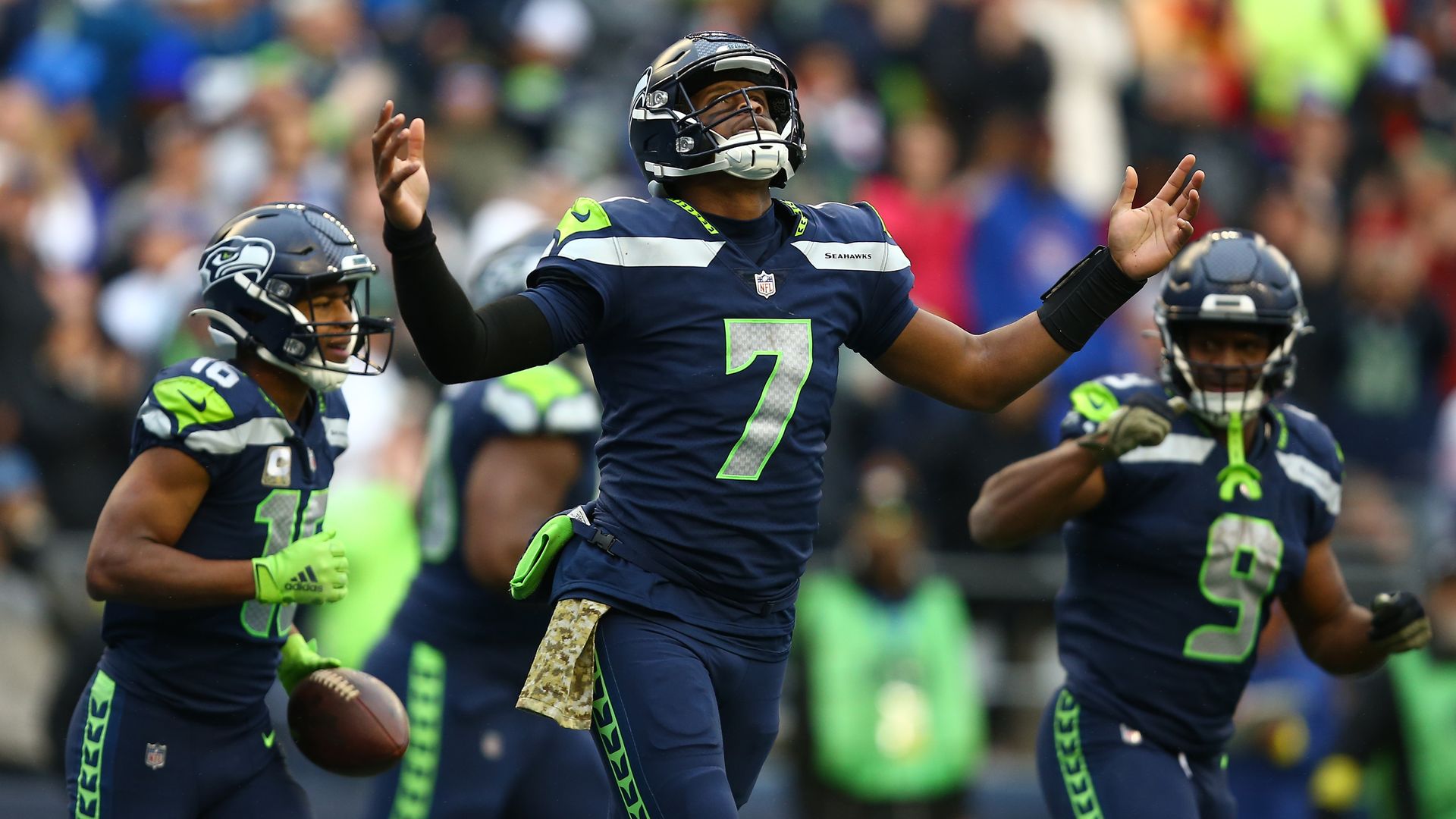A football player in a dark blue jersey and the number 7 holds his hands up in a quiet celebration on a football field.