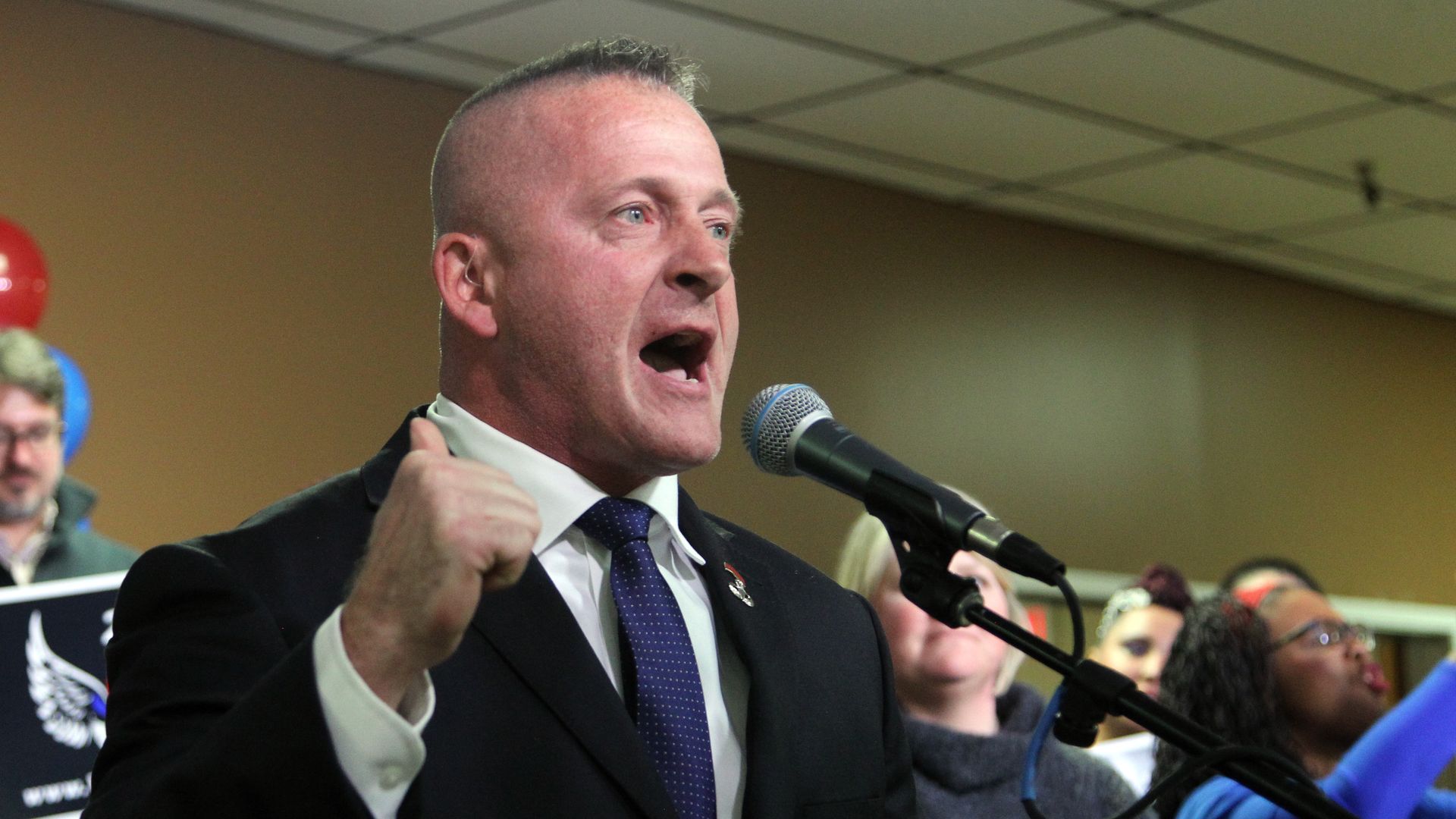 Richard Ojeda, wearing a dark gray suit and speaking into a microphone in front of a wood-paneled room while surrounded by supporters.