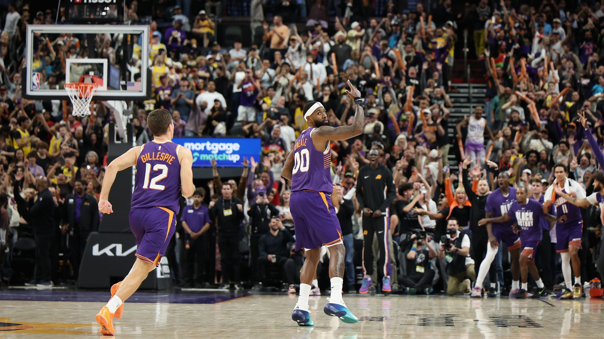 A basketball player in a purple uniform gestures in the air with his right hand while a teammate runs toward him in front of a packed arena.