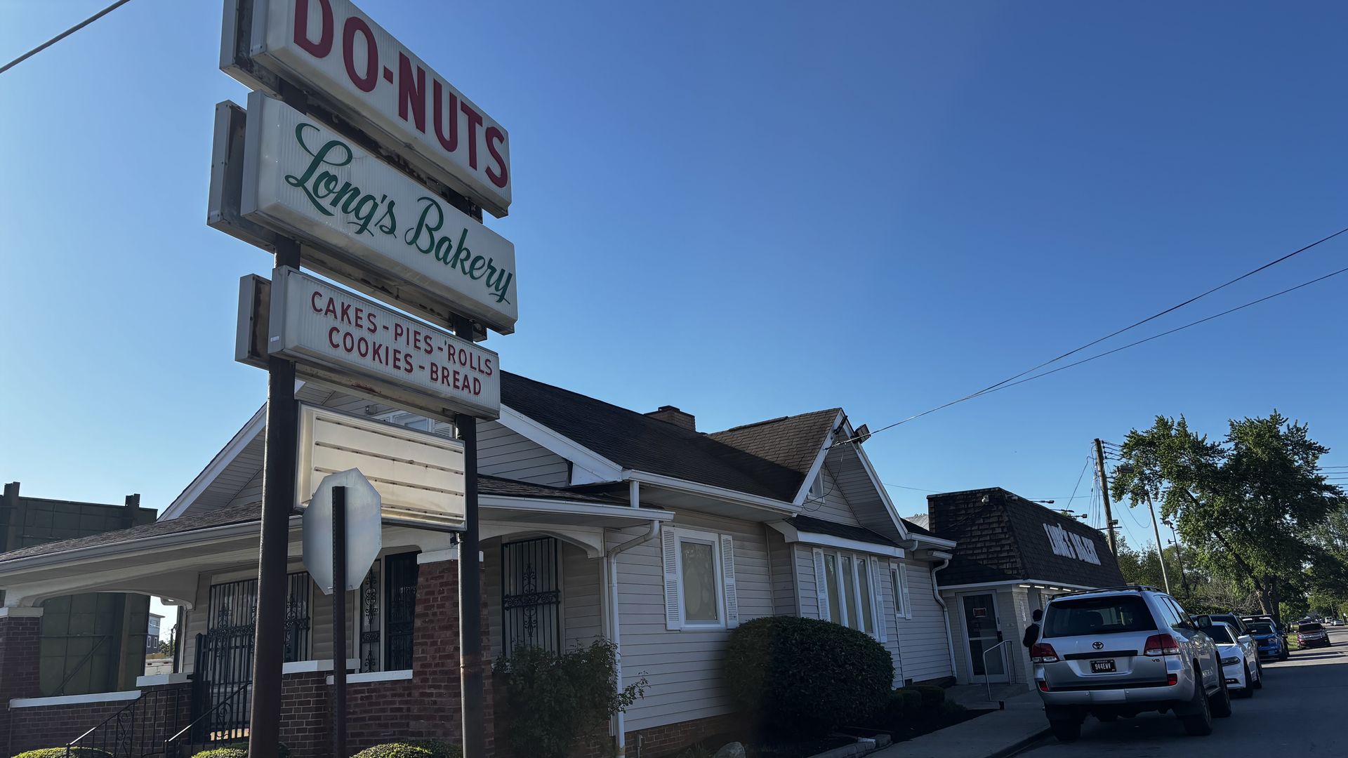Sunny street with a tall three-panel bakery sign: red "DONUTS" on top, green script "Bakery" middle, red "CAKES-PIES-ROLLS COOKIES-BREAD" bottom; blue sky and houses in background.