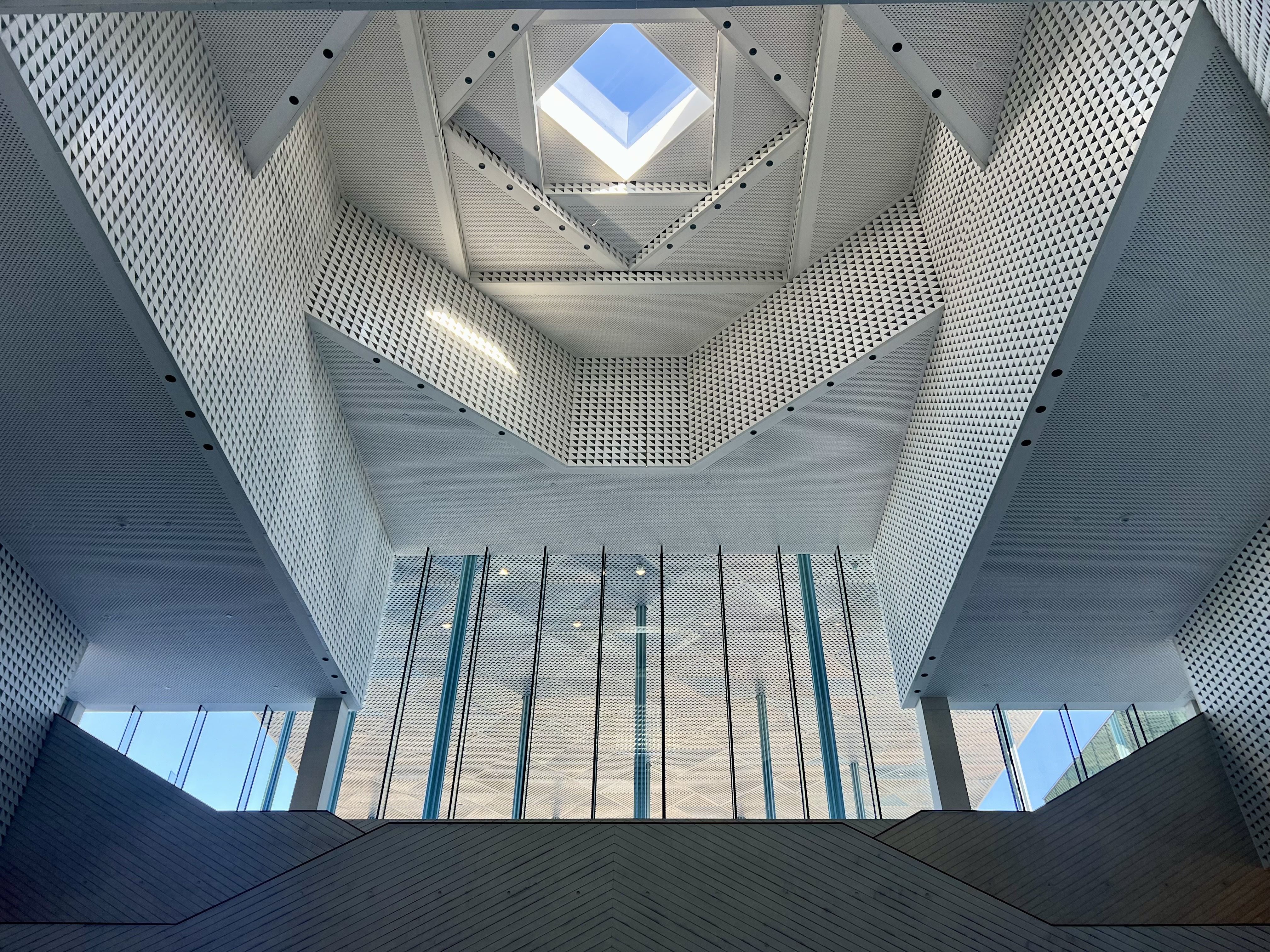 Interior view of a modern building with geometric white patterned walls, vertical glass panels, and a skylight at the center letting in blue sky light.