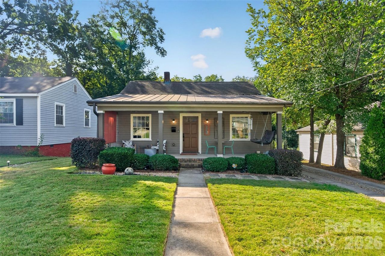 Single-story gray house with a covered front porch, white columns, and a brown door. Turquoise chairs and a porch swing sit on the porch; green lawn, concrete path, a neighboring blue house, and trees in the background.