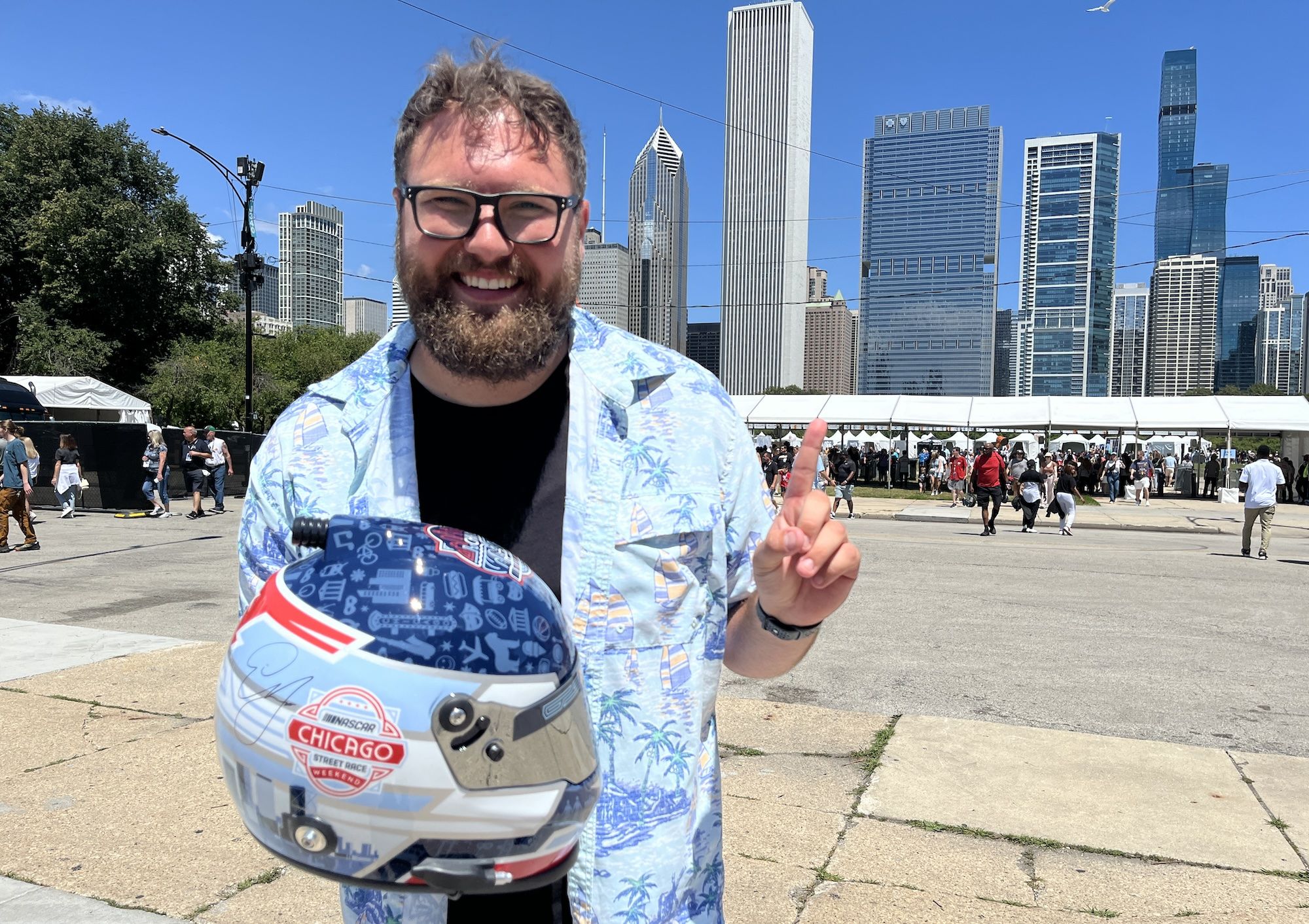 Man in blue shirt holds a racecar helmet, holds up a finger indicating #1 and stands in front of Chicago skyline.