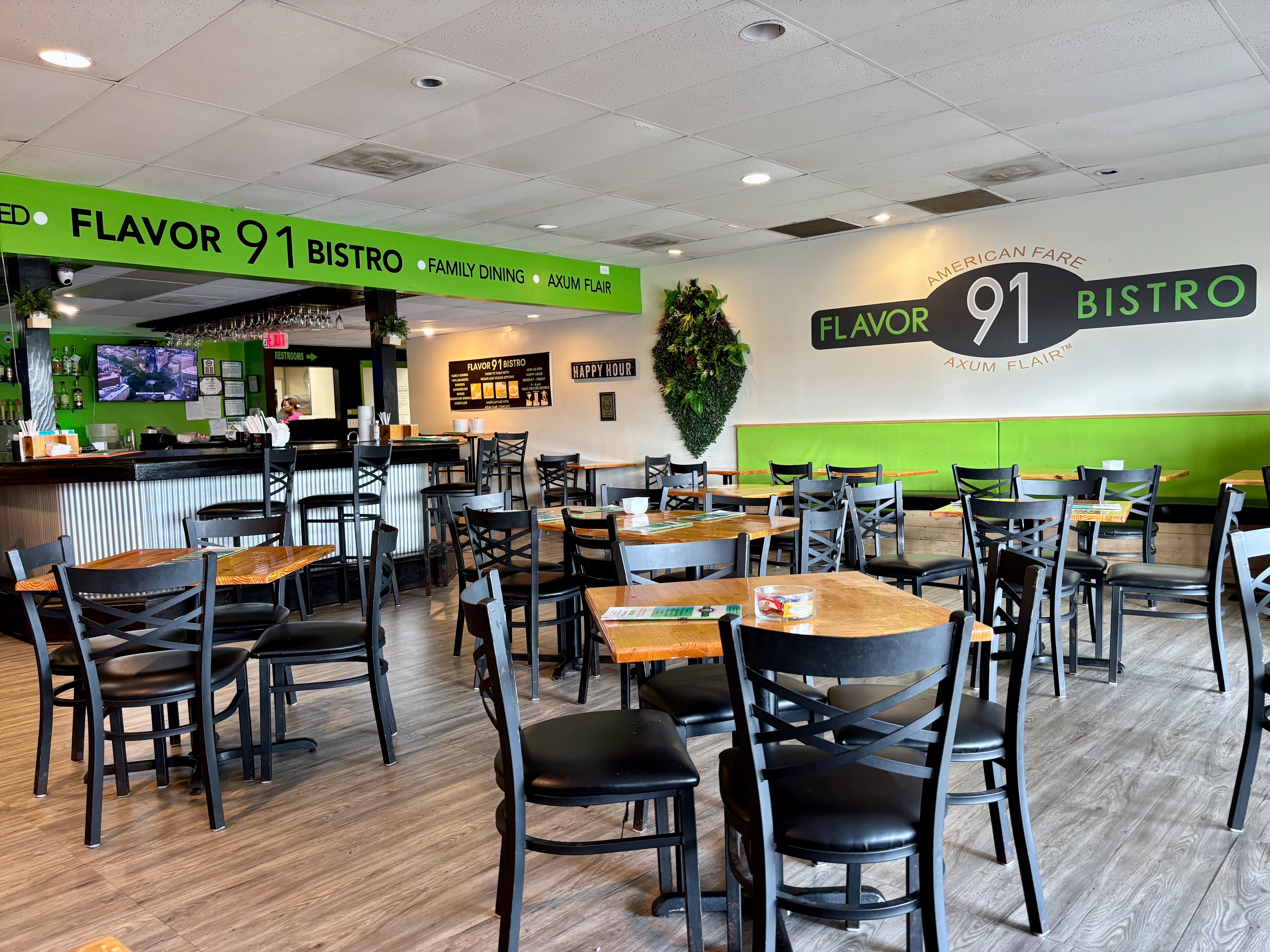 Interior of Flavor 91 Bistro with black chairs, wooden tables, green accents, bar area, a TVs, and signs featuring "Flavor 91 Bistro," family dining, and happy hour details.