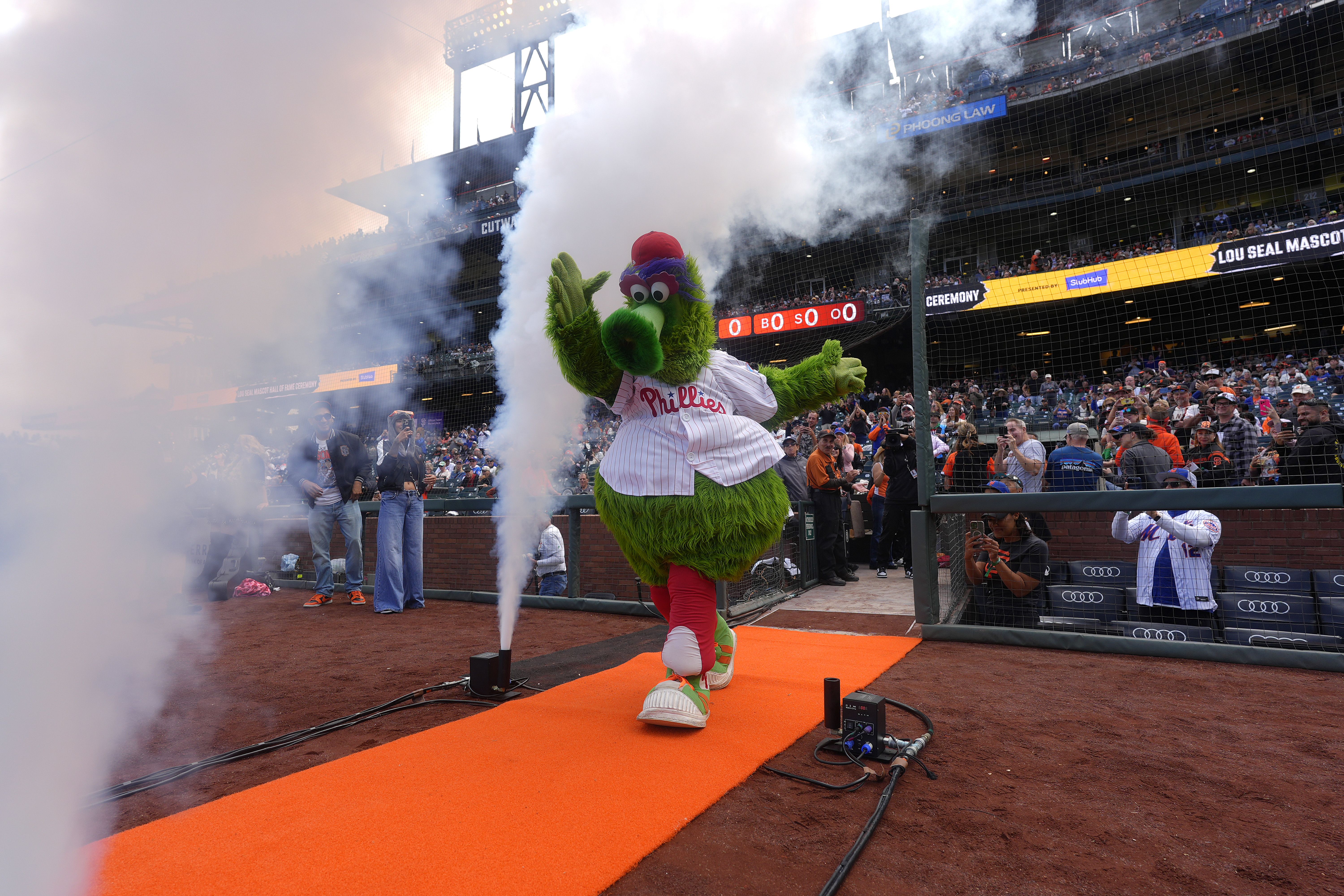 Philadelphia Phillies mascot wearing red cap and white pinstripe jersey walks down orange carpet with smoke effects, fans watch and take photos at baseball stadium event.