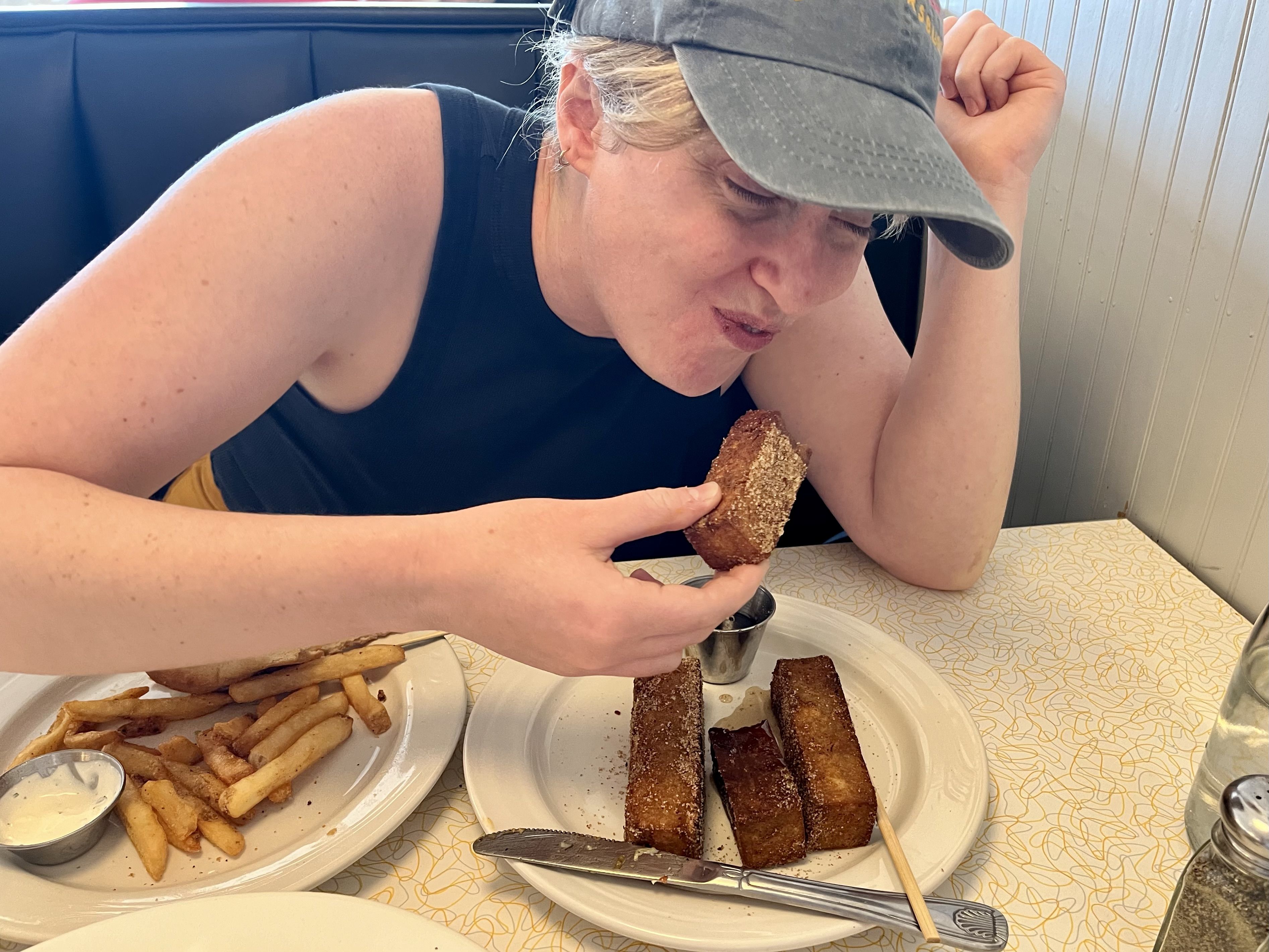 A woman in a black top and blue baseball cap smiling after taking a bite of a French toast stick.